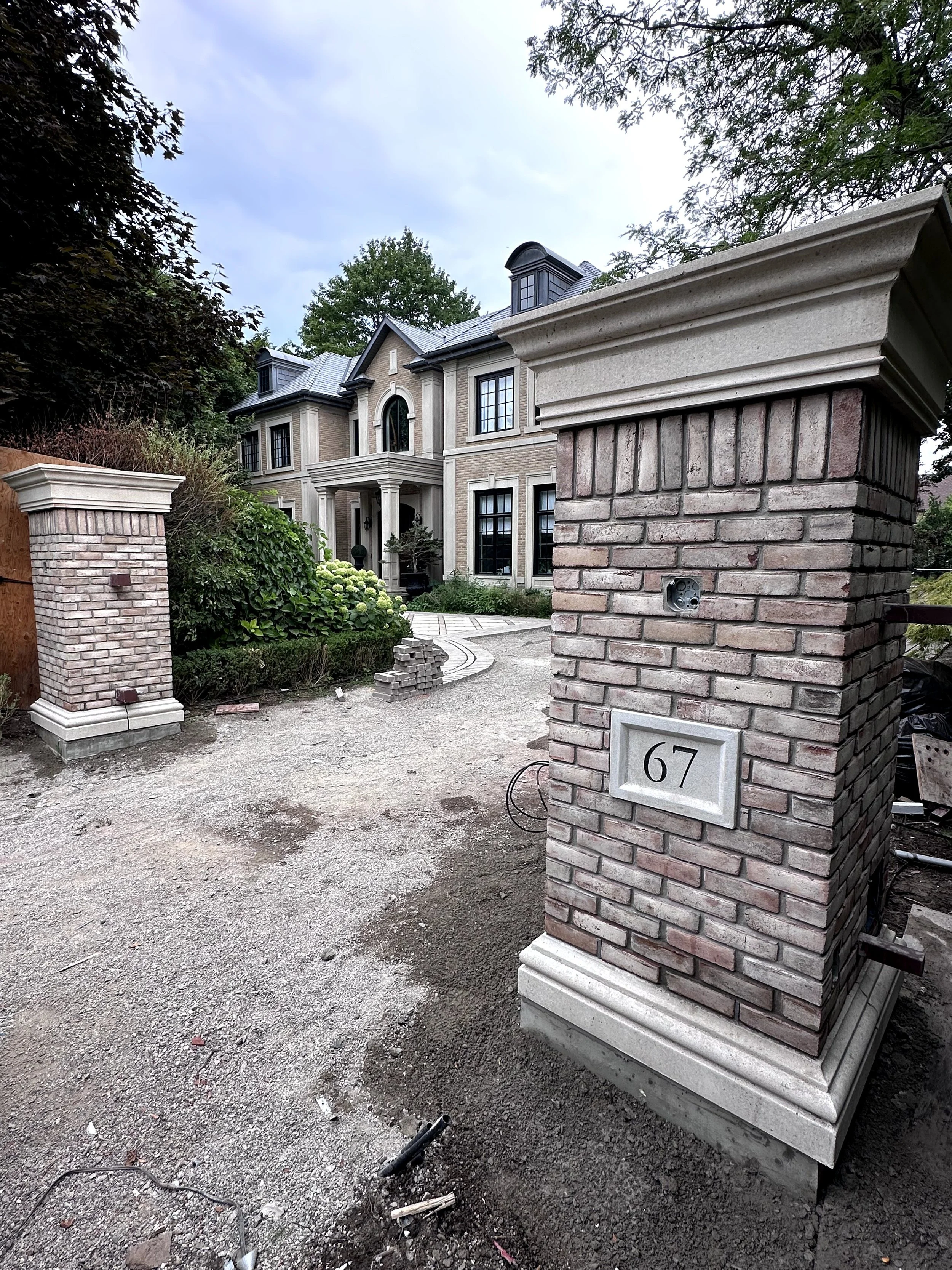 A large brick house with columns, multiple windows, and a garage, behind a brick letterbox with the house number 67, under a cloudy sky. There is landscaping with trees and bushes surrounding the house.