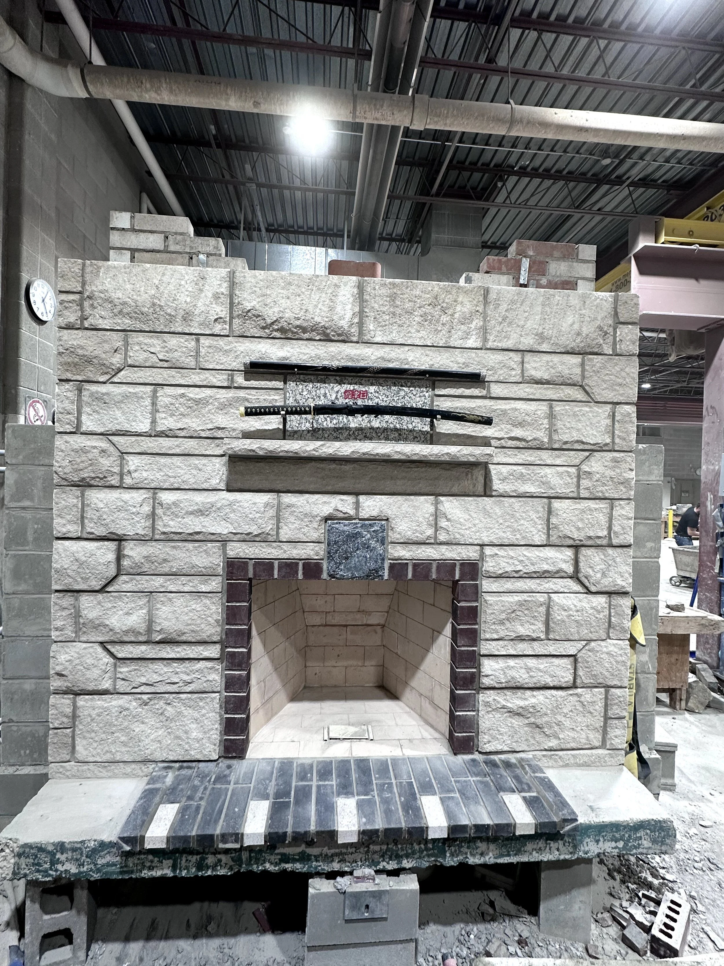 A stone fireplace under construction with a sword and a katana displayed on a wooden platform in front of it, surrounded by construction tools and materials.