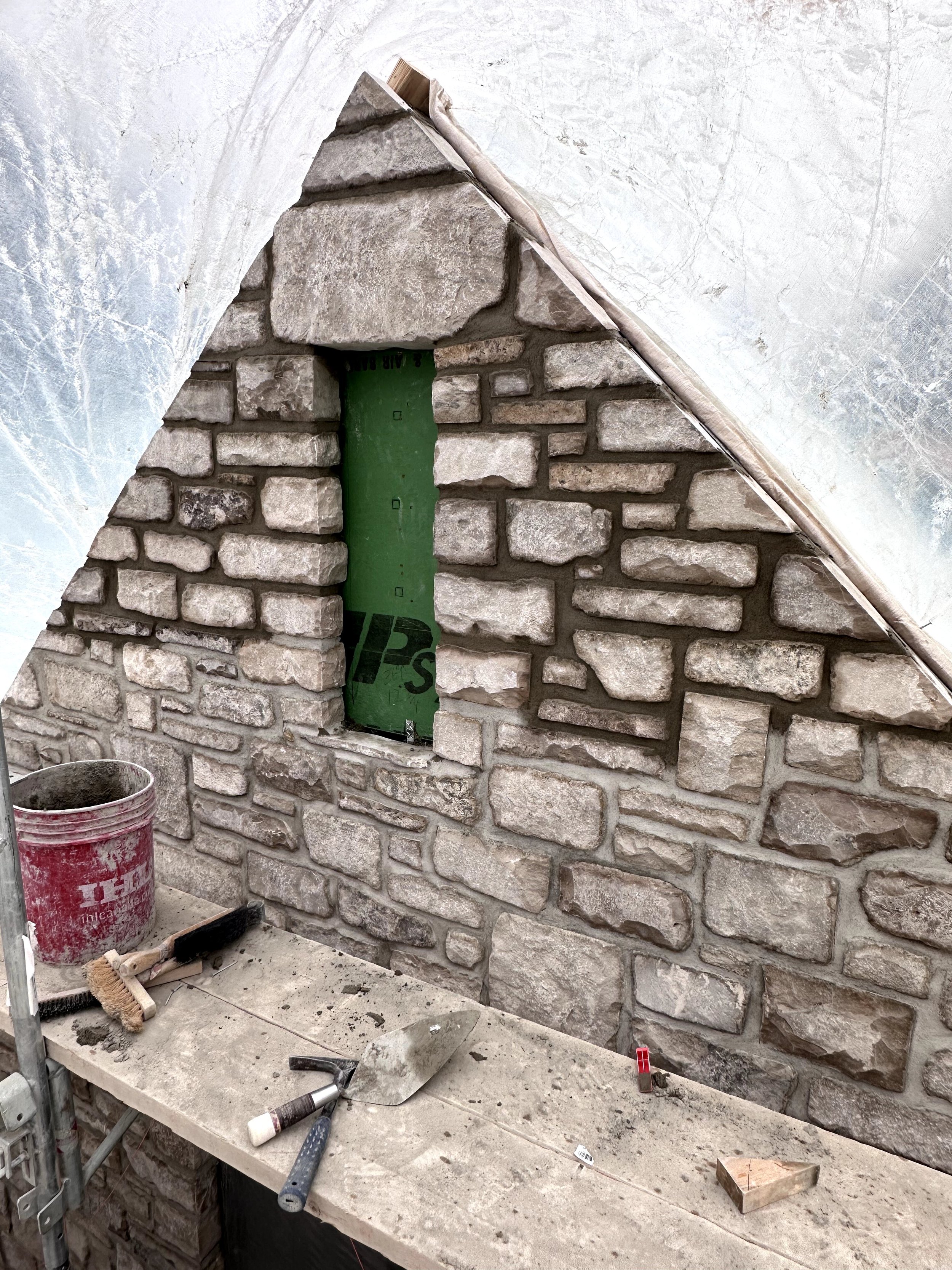 Interior of a construction site with a partially built stone wall, a workbench with various tools, a bucket, and construction materials.