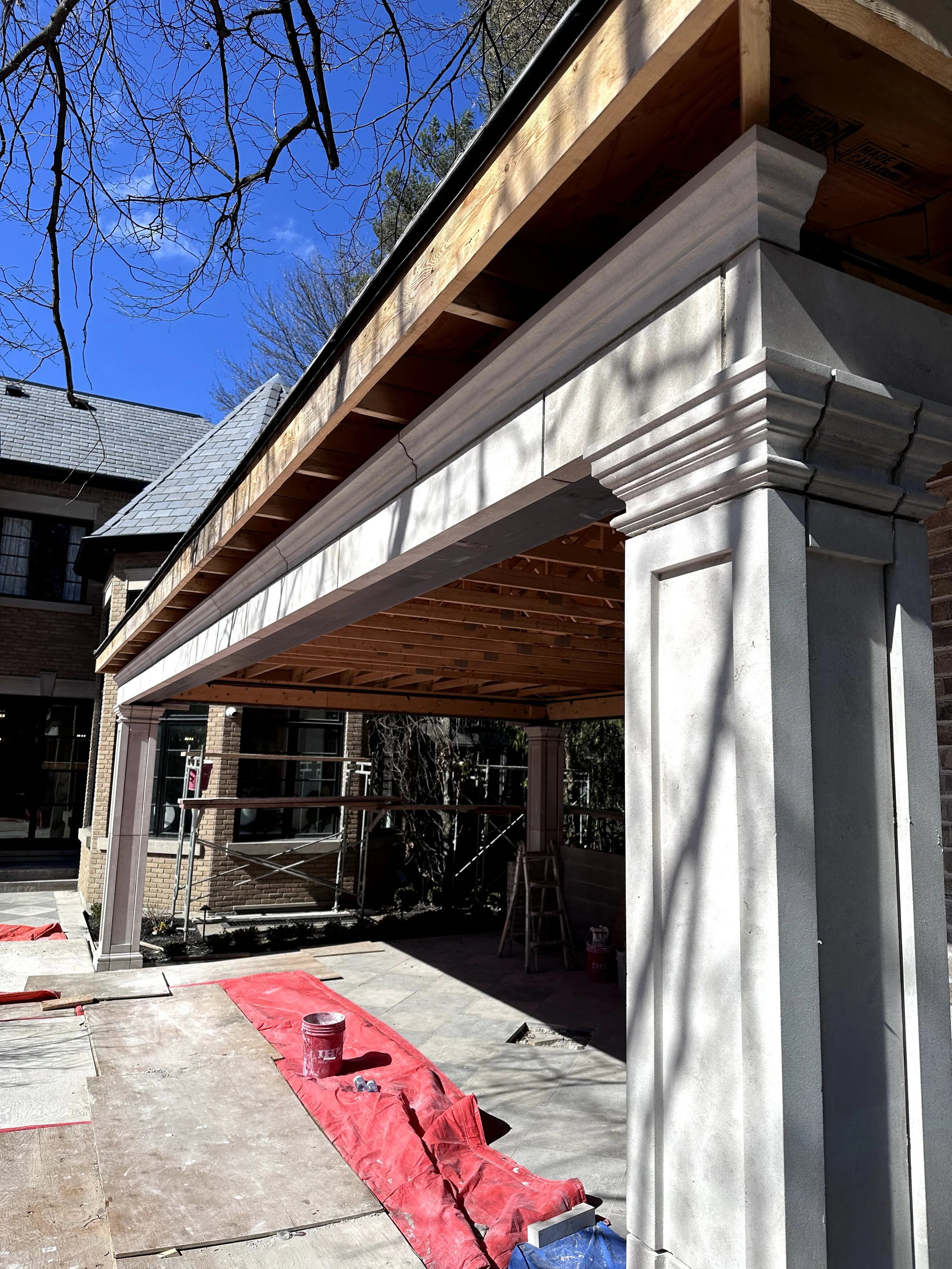 Construction site showing a building with a brick wall, a concrete column with decorative molding, and an overhang with wooden framing. Construction materials and tools are on the ground, and scaffolding is visible in the background. The sky is clear