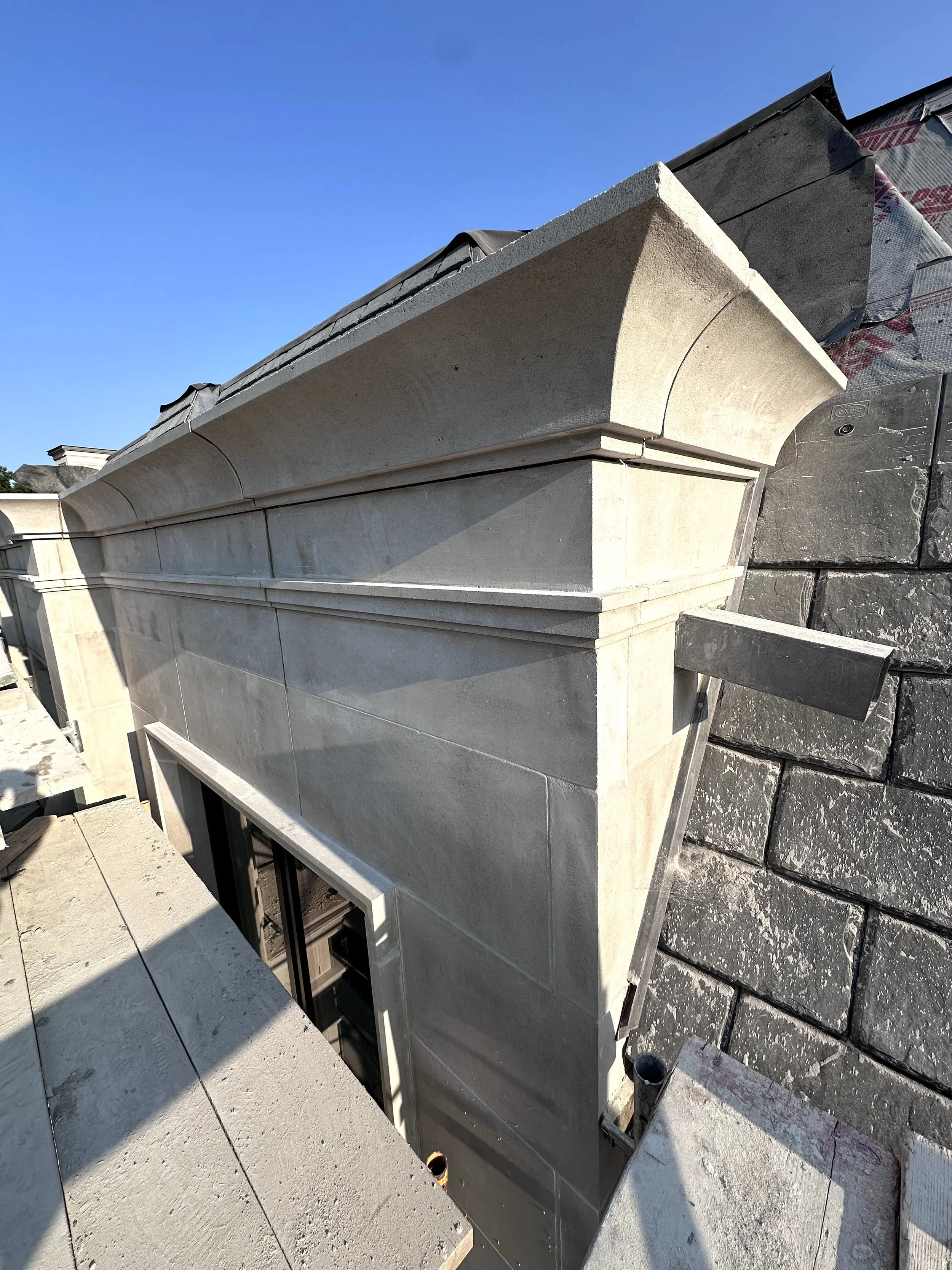 Construction site showing the exterior of a building with stone siding, a roof, and a small window, with construction materials and scaffolding around.