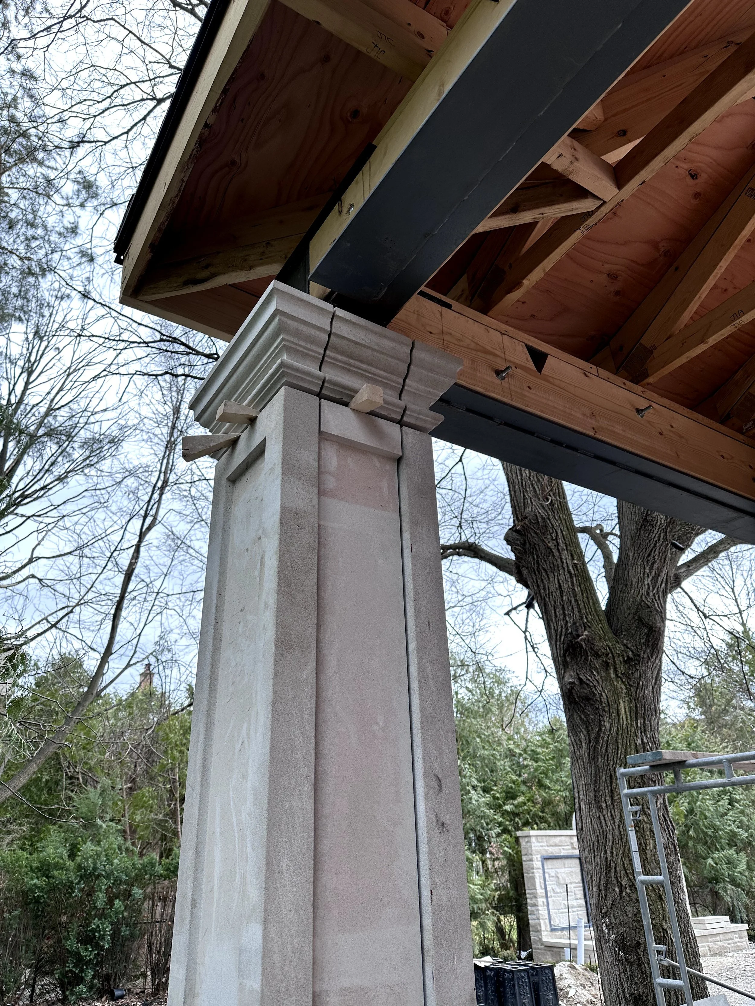 Under construction outdoor structure with a stone column, wooden beams, and a black metal support beam. There is a ladder on the right side, trees in the background, and construction materials on the ground.