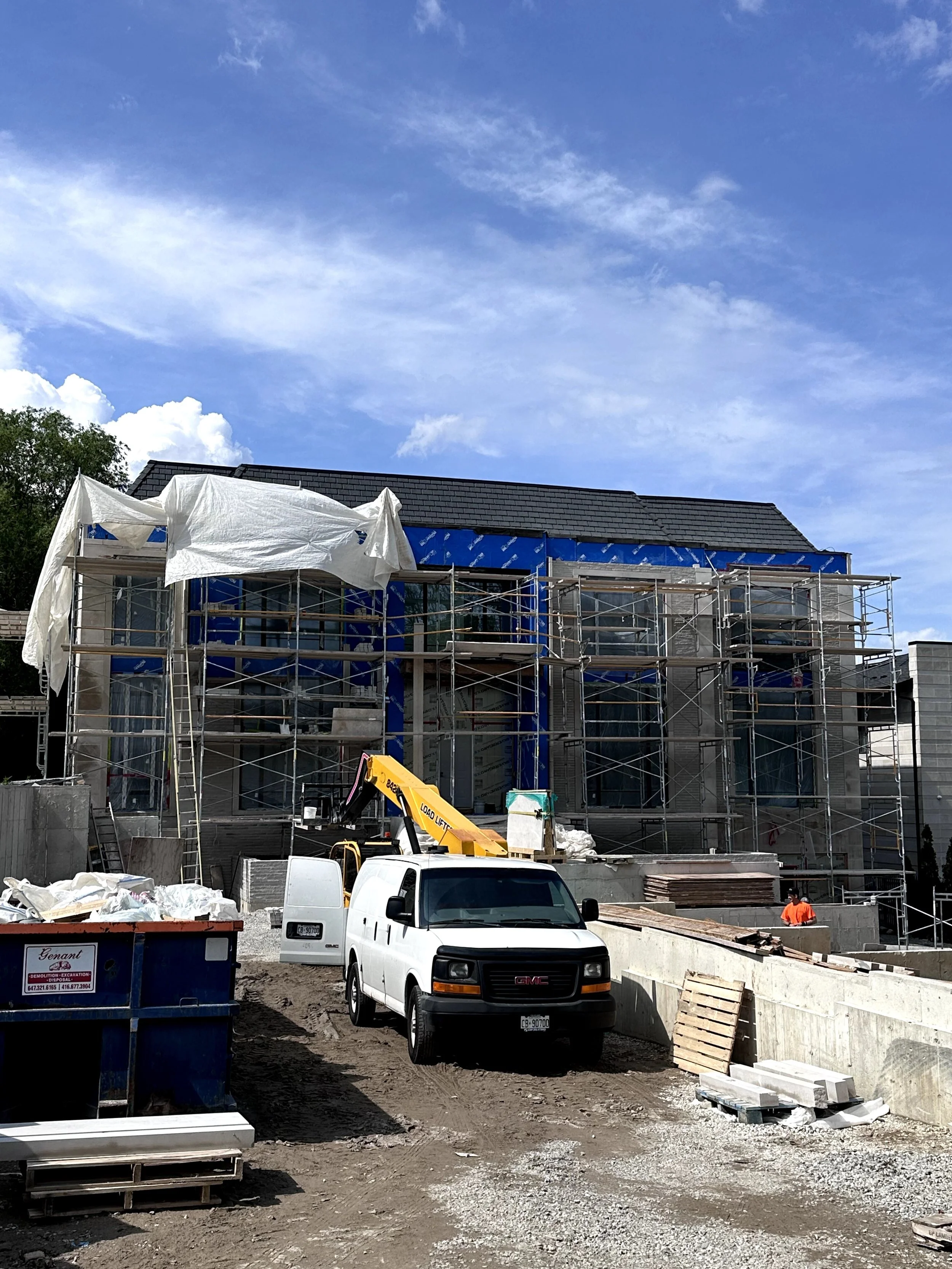 Construction site with scaffolding around a multi-story building, construction vehicles, and workers, with a blue sky and clouds.