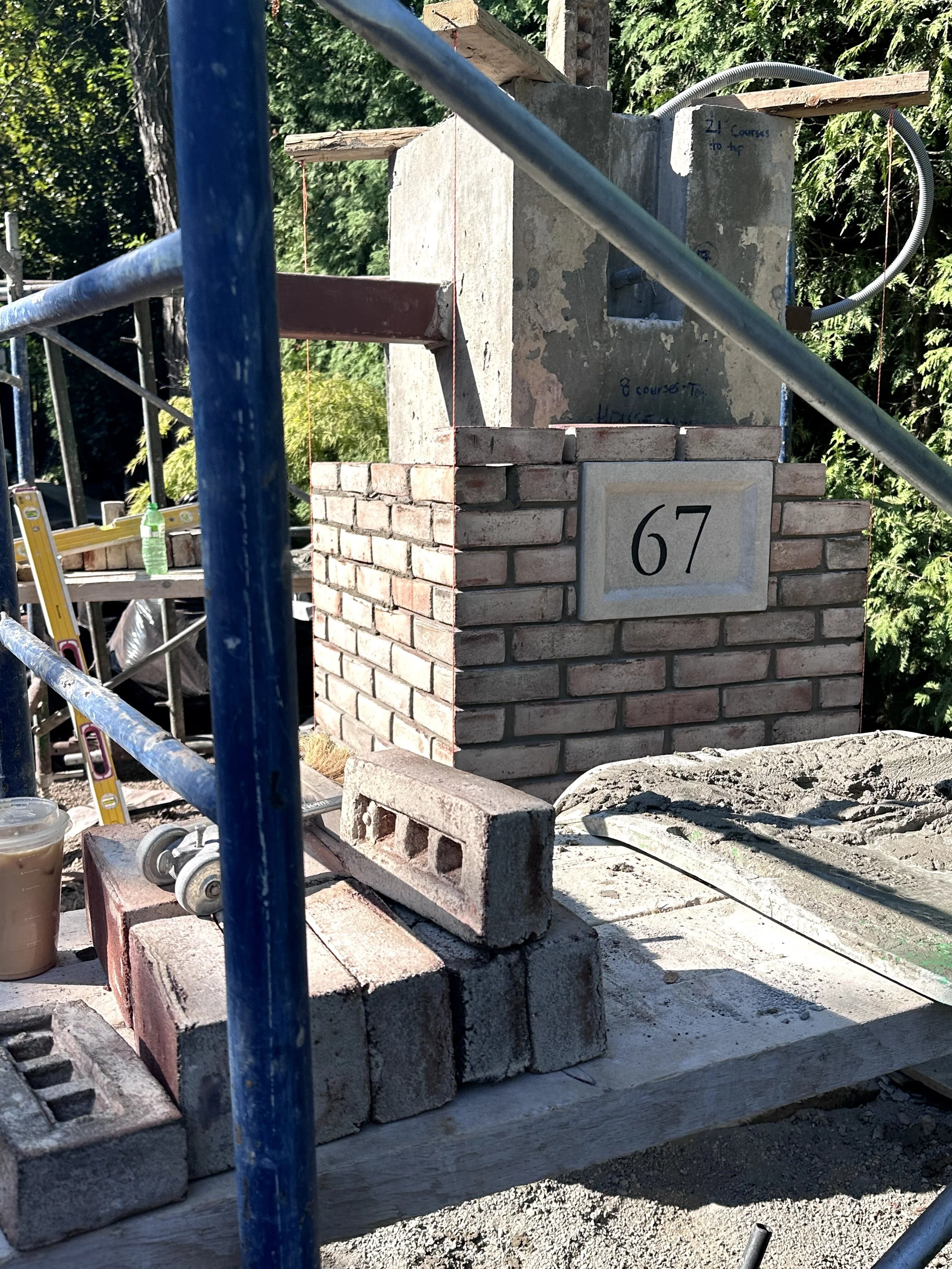 Under construction brick chimney with a concrete cap, surrounded by scaffolding, construction tools, and a bottle of water, with trees in the background.