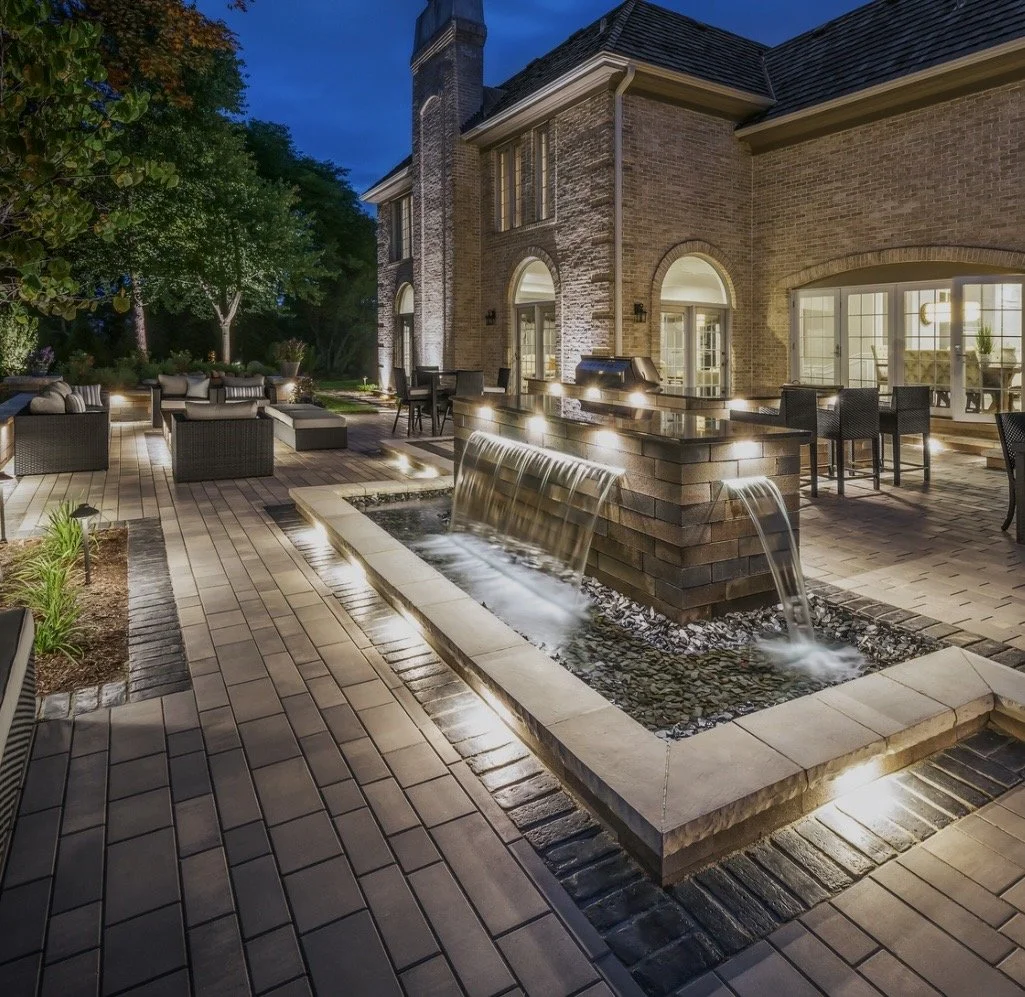 Nighttime view of an elegant backyard patio with a brick house, illuminated water fountain, outdoor seating, and lush trees.