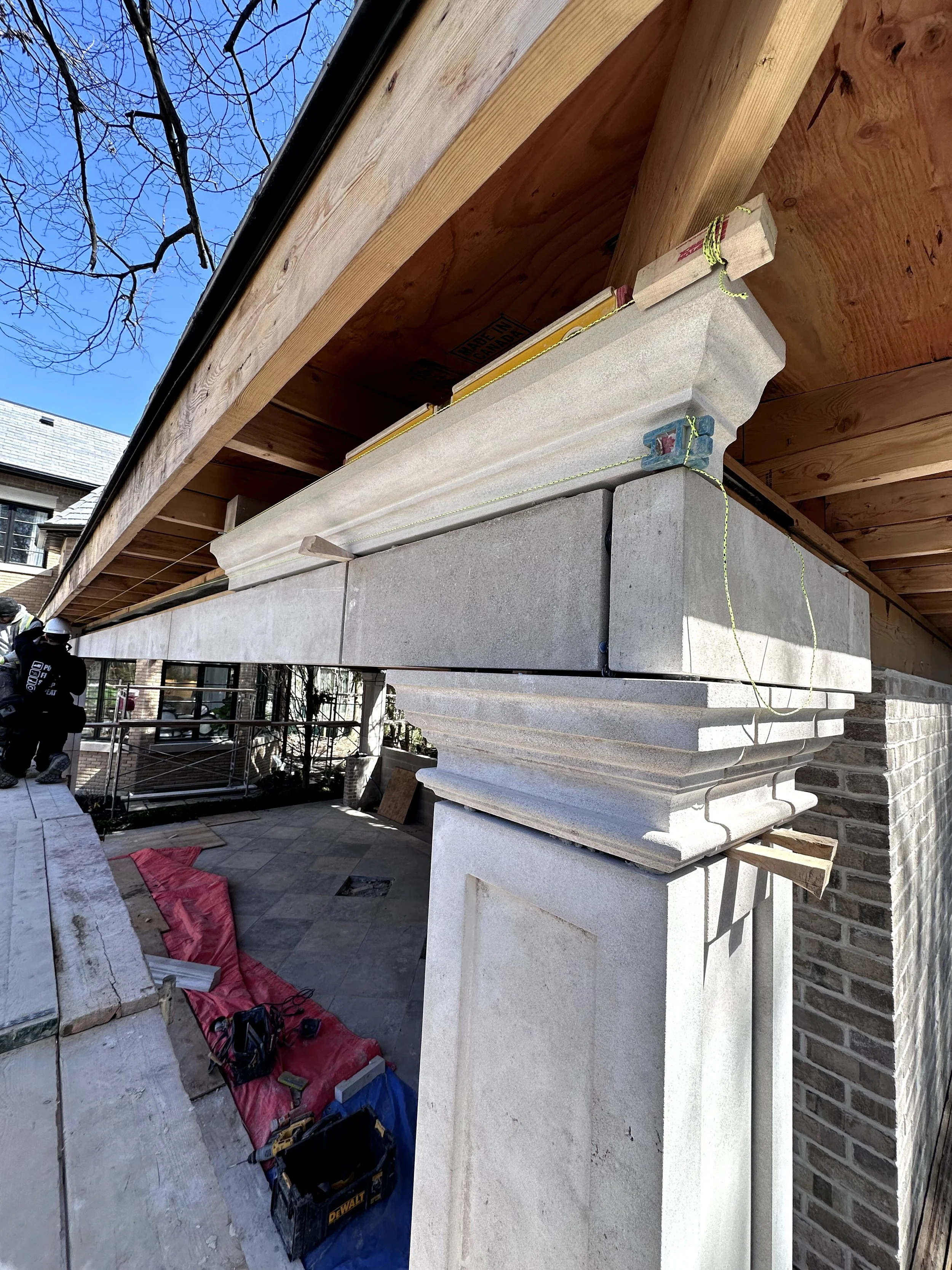 Construction workers installing large stone columns on the porch of a house with brick and wooden exterior. Tools and construction materials are scattered on the ground.