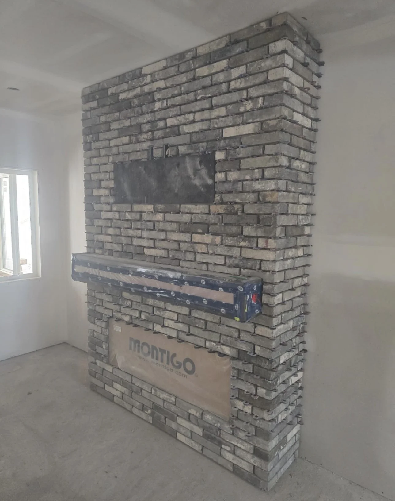Interior of a room with a partially built brick fireplace and a window on the left wall, construction materials, and an unfinished concrete floor.