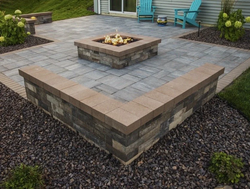 Raised brick fire pit on a paved patio with Adirondack chairs and landscaping in the background.