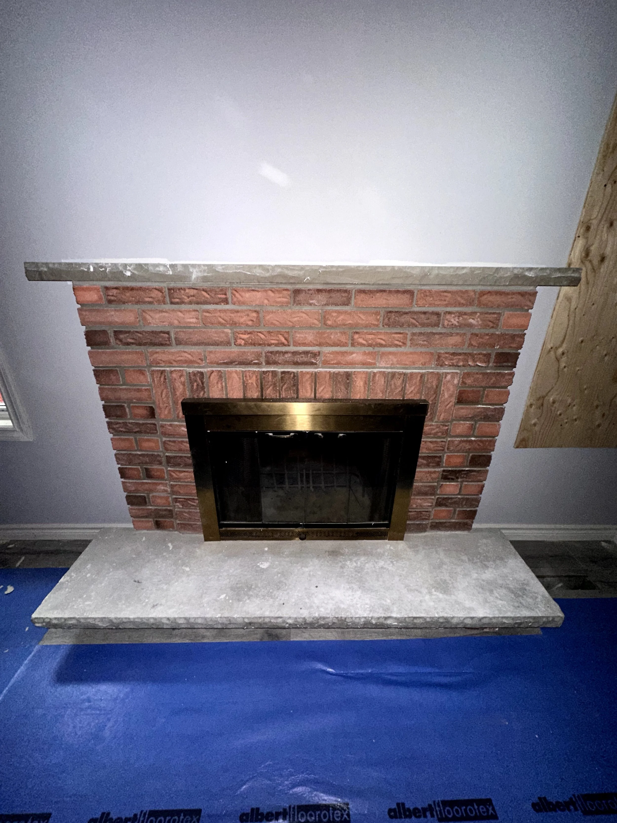 A brick fireplace with a concrete mantel, framed by a gold trim on the opening and surrounded by white walls and a wooden beam on the right.