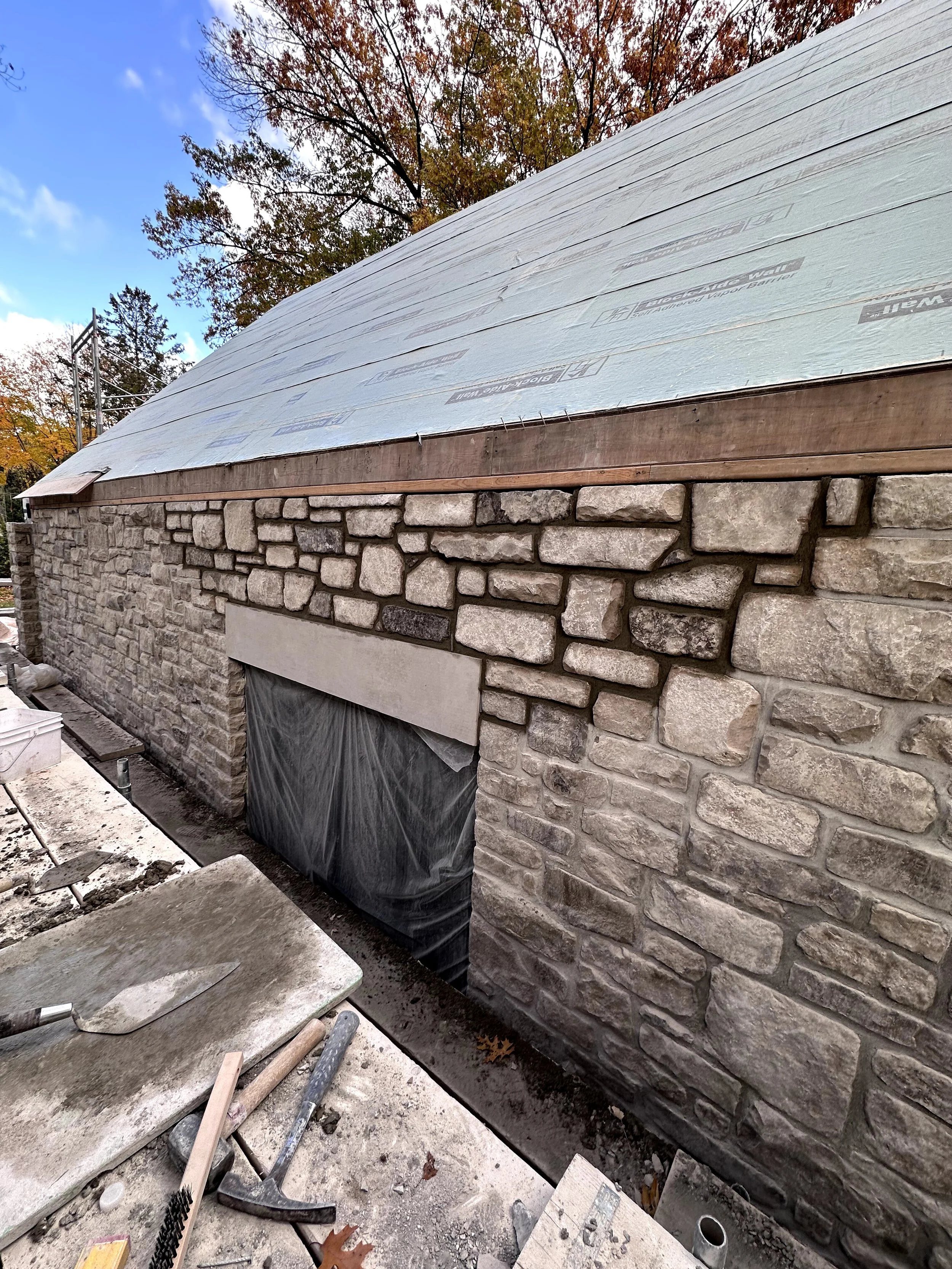 Construction site with stone wall and building under renovation, scaffolding visible in the background, tools on the ground, and a partially finished stone exterior wall.