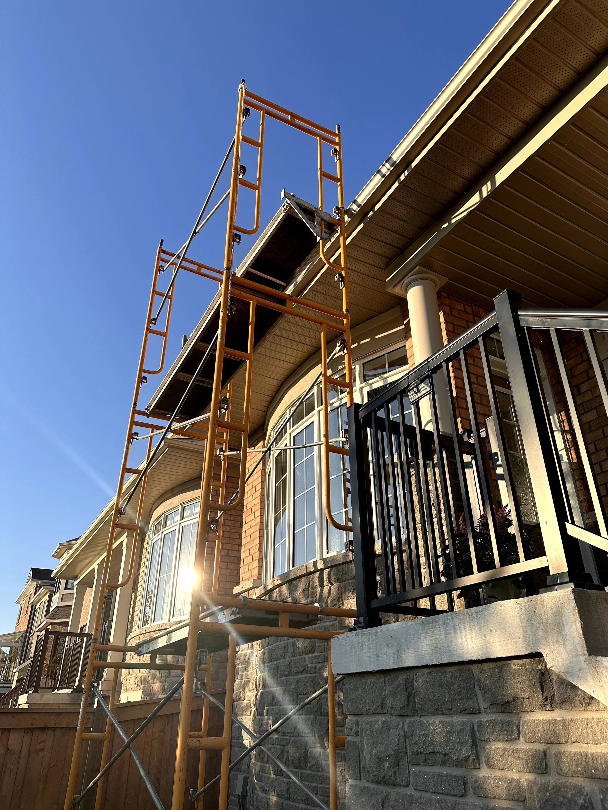 Scaffolding set up along the exterior of a brick house, with large windows and a black metal gate in the foreground, under a clear blue sky.