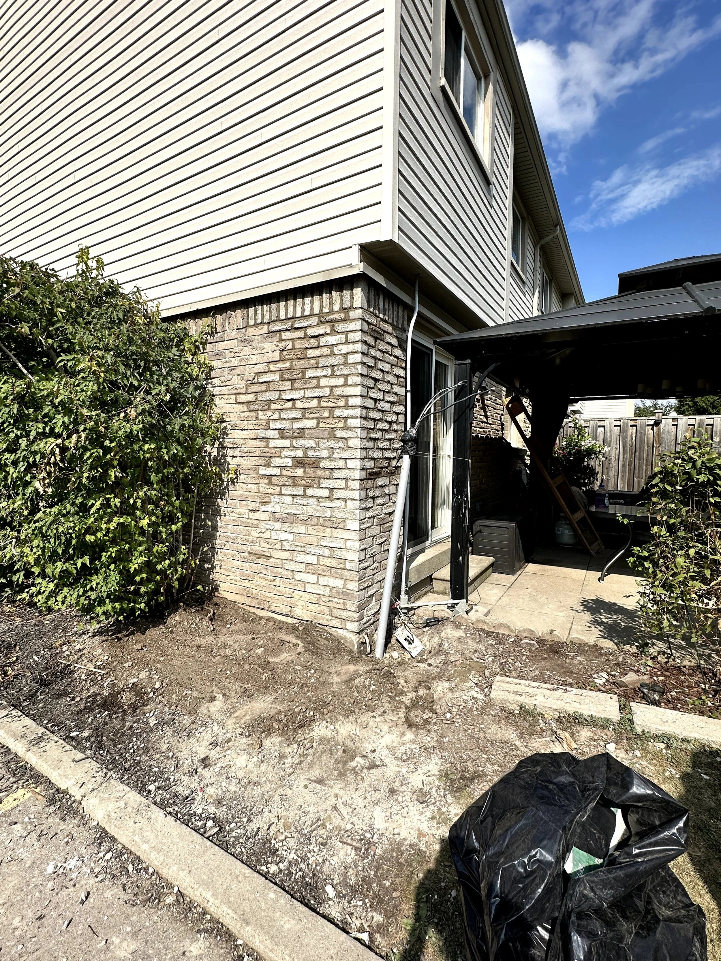 Exterior view of a house with a brick and siding wall, a sliding glass door, a plant, and a covered area with gardening tools. The sky is partly cloudy.