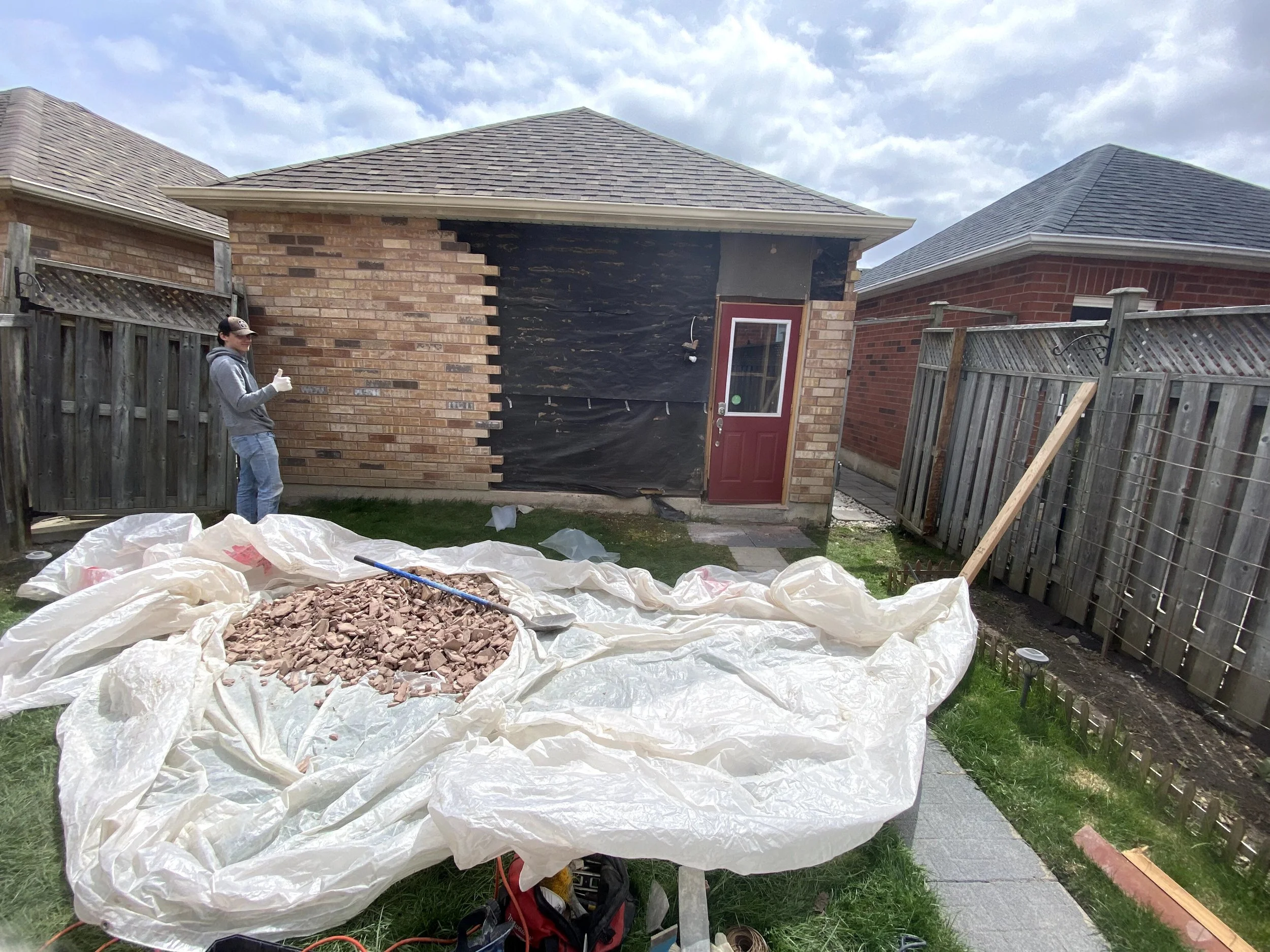 A person standing next to a wooden fence giving a thumbs-up, while a pile of broken bricks covered with a plastic sheet is in the foreground. The building in the background has partially removed siding revealing black tar paper underneath. The scene 
