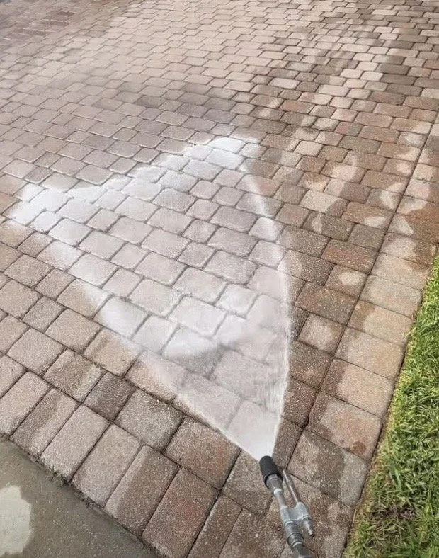 A person uses a pressure washer to clean a brick walkway, removing dirt and grime and revealing cleaner bricks underneath.