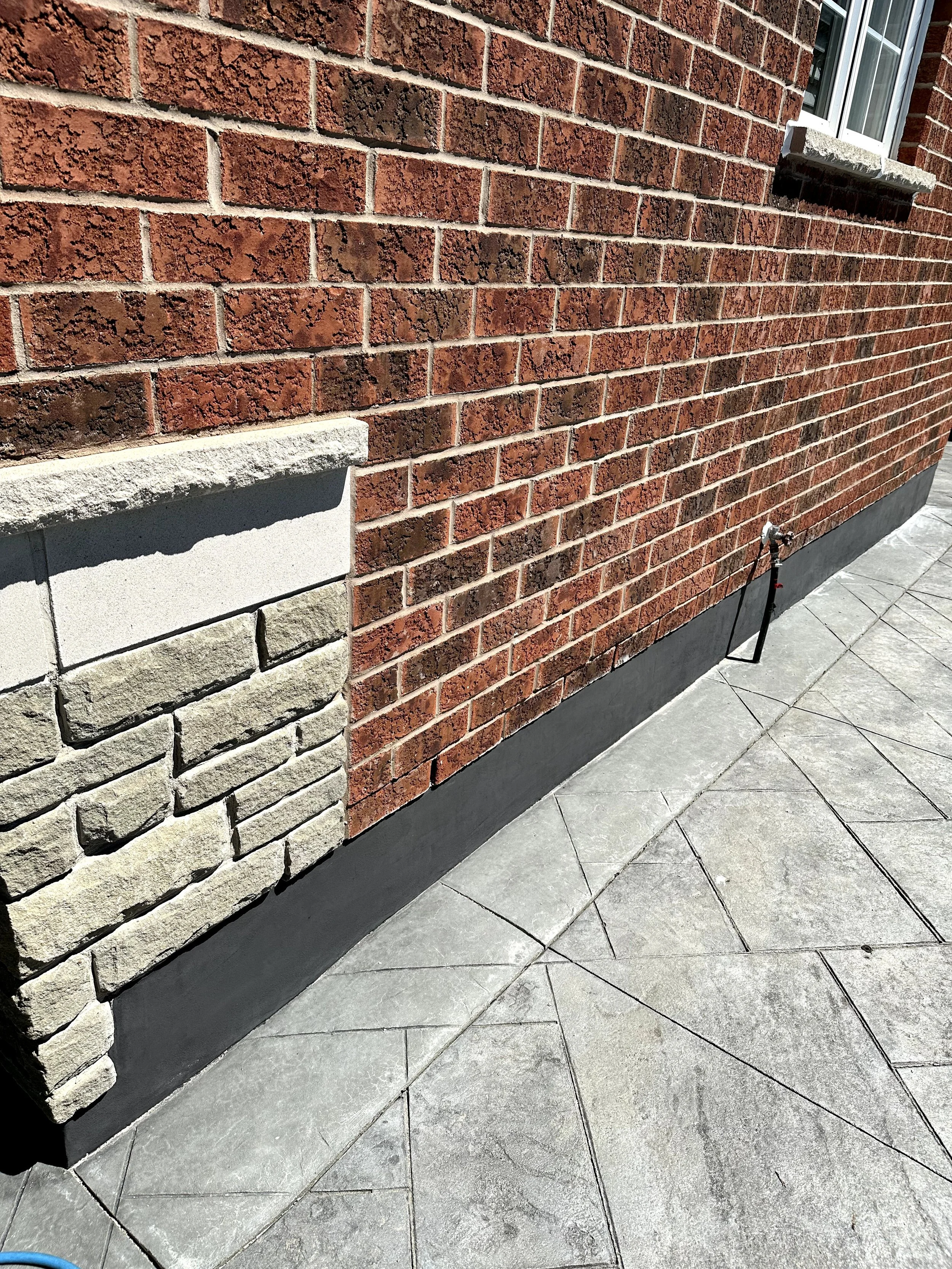 Close-up of a brick wall with a decorative stone corner and a small window with a brick sill. The ground is made of gray stone tiles.