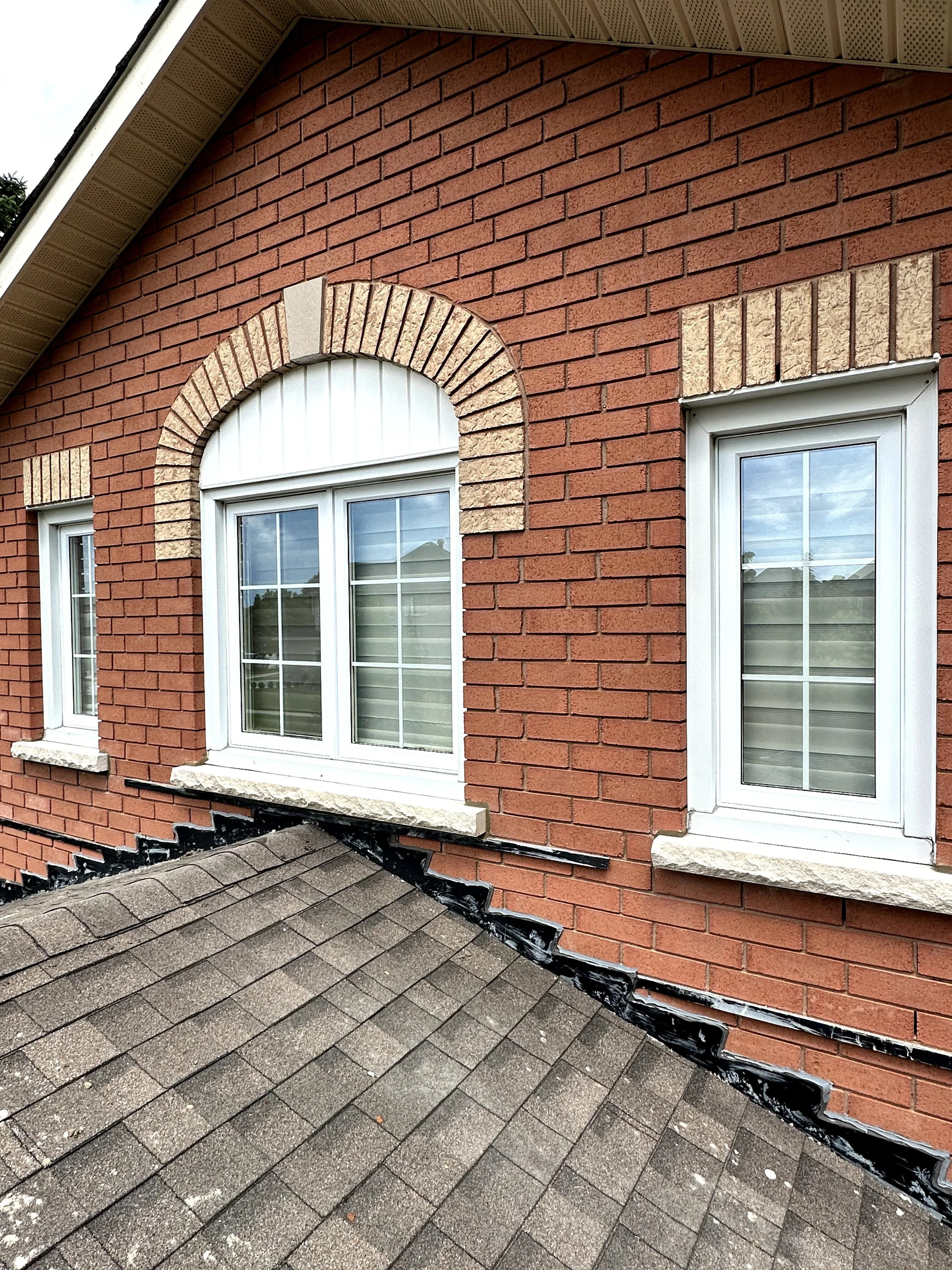 Close-up of a brick house exterior with two white-framed windows. The bricks are a reddish-brown color, with decorative beige brick accents above the windows. The roof has gray shingles, and there's a section of the roof's edge and gutter visible. Bl