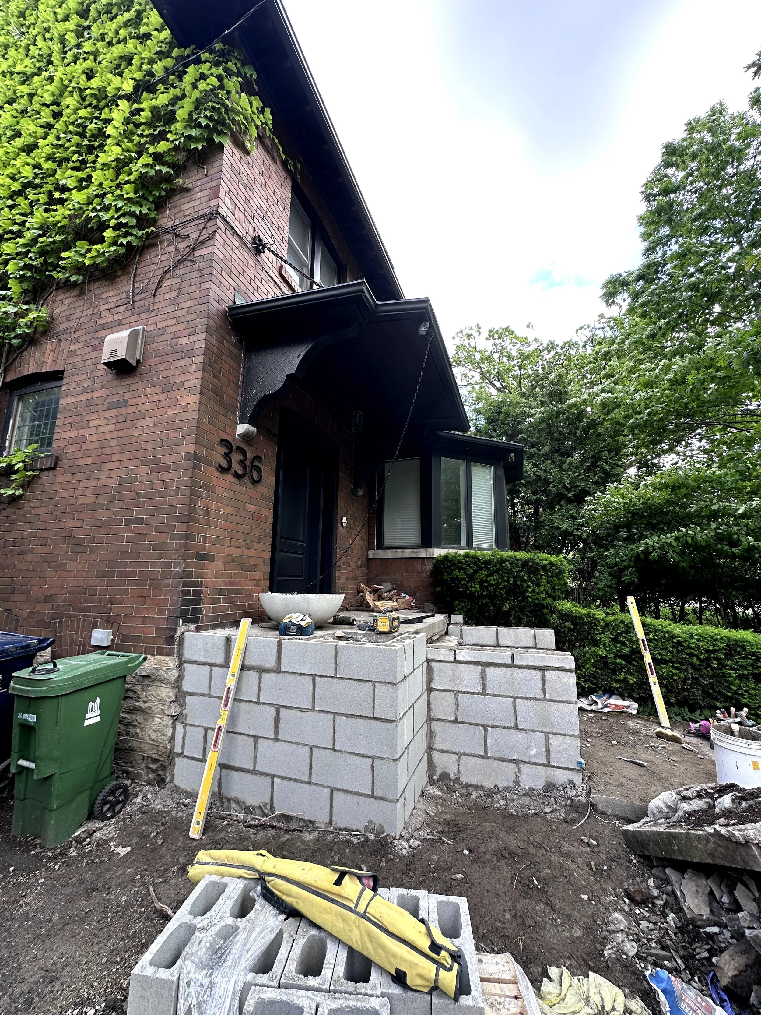 Construction site in front of a brick house, with concrete blocks, tools, and construction materials, and trees in the background.