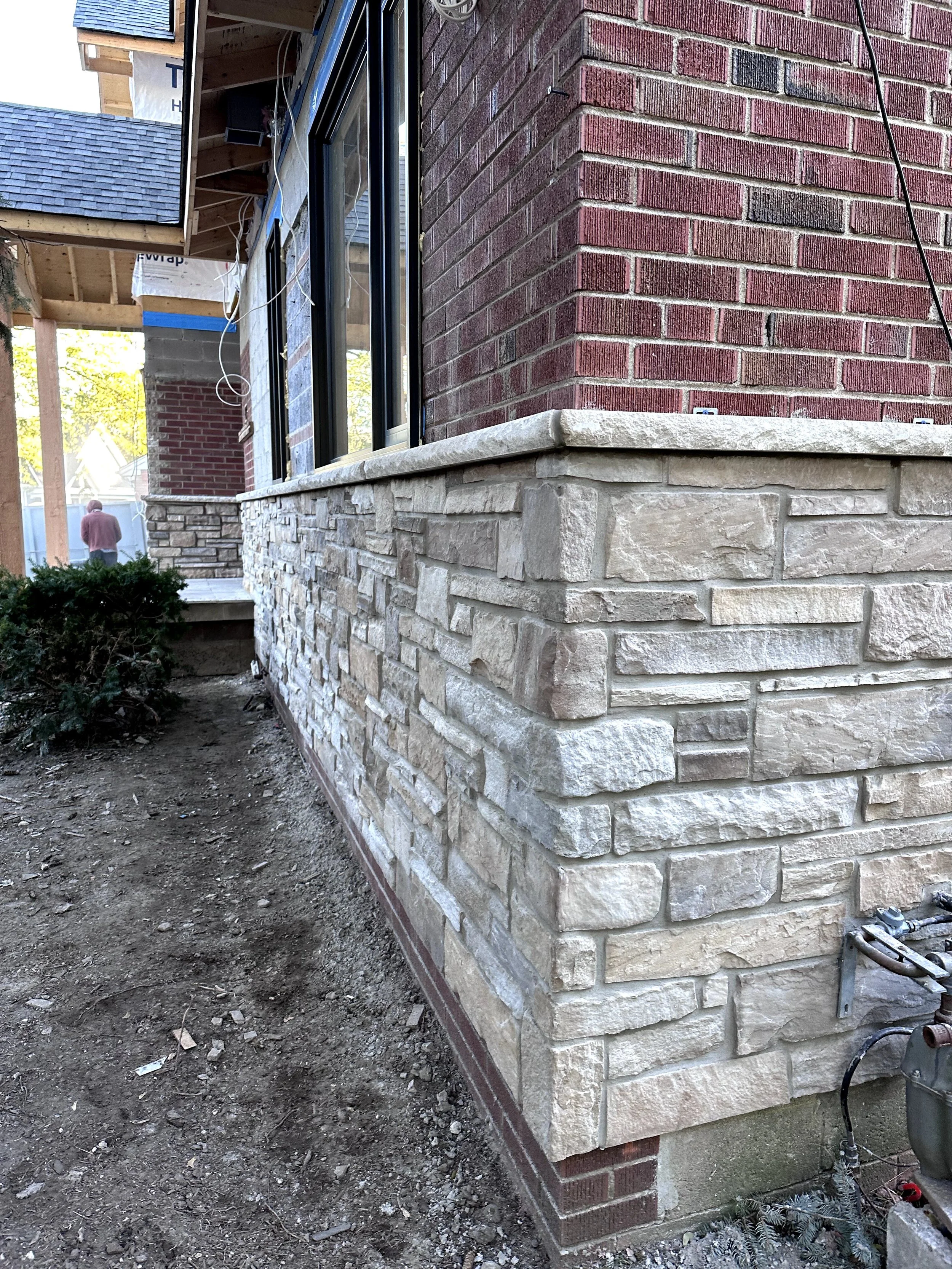 A house under construction with a brick exterior wall, a stone foundation, and a large window. There is construction material and equipment nearby, with soil and debris on the ground.