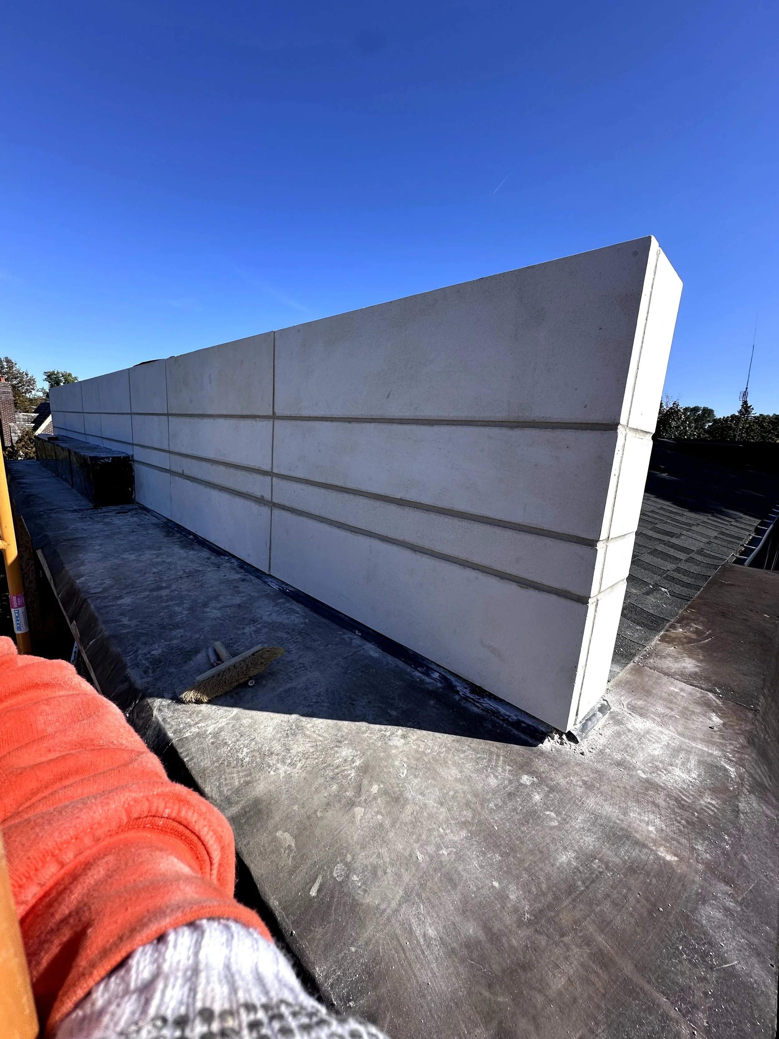 View of a rooftop with a row of large, white concrete blocks against a clear blue sky. Part of a person's arm in a red jacket and gray and white sock is visible in the foreground.