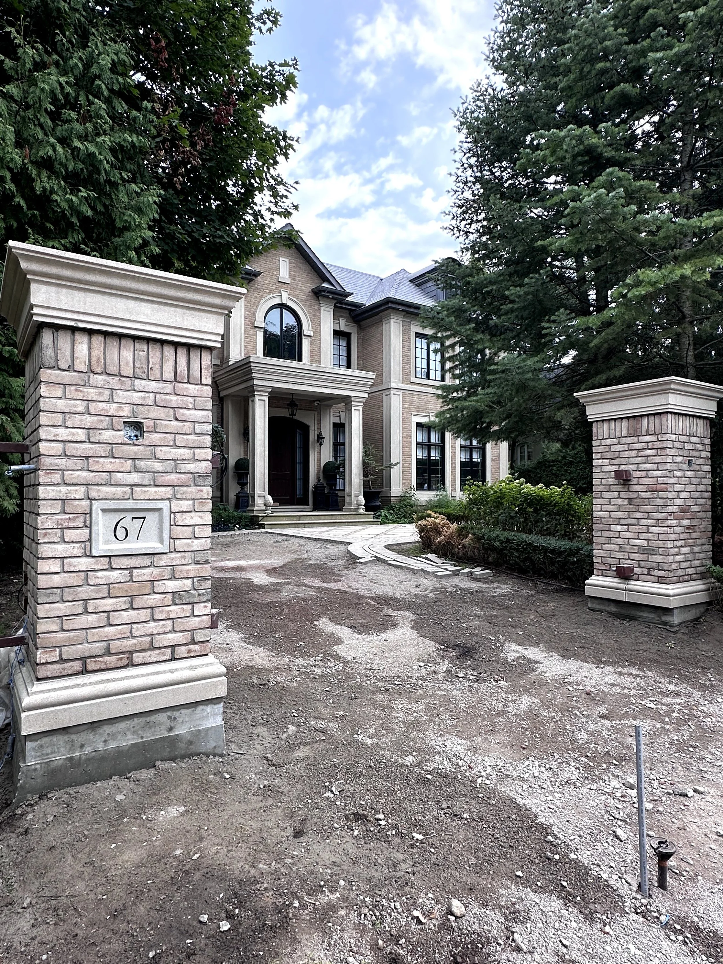 Front of a large house with a brick and stone facade, large arched window, and columns at the entrance, surrounded by trees and a brick gate with the house number 67.