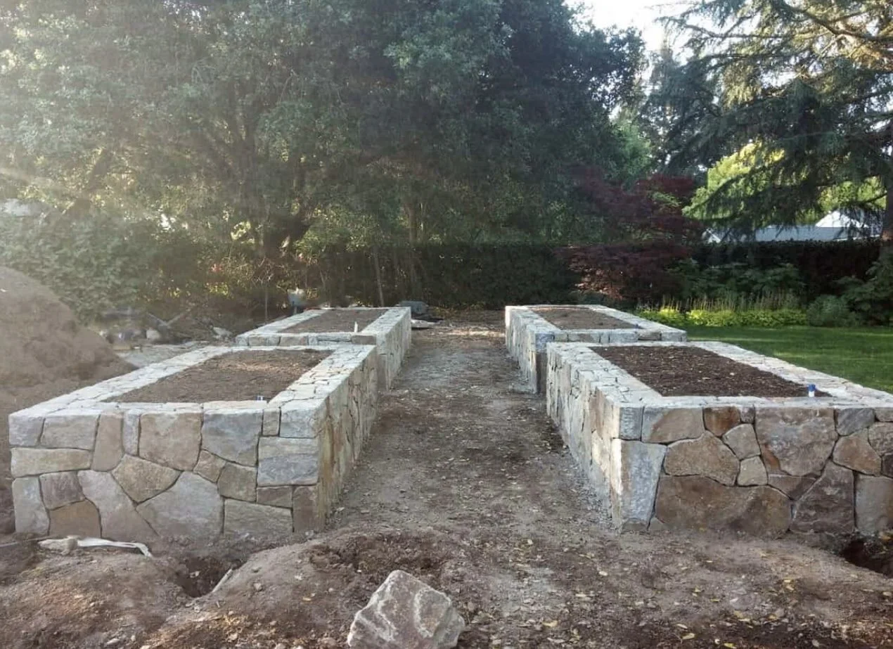 Stone garden beds under construction with soil, in a backyard with trees and grass in the background.