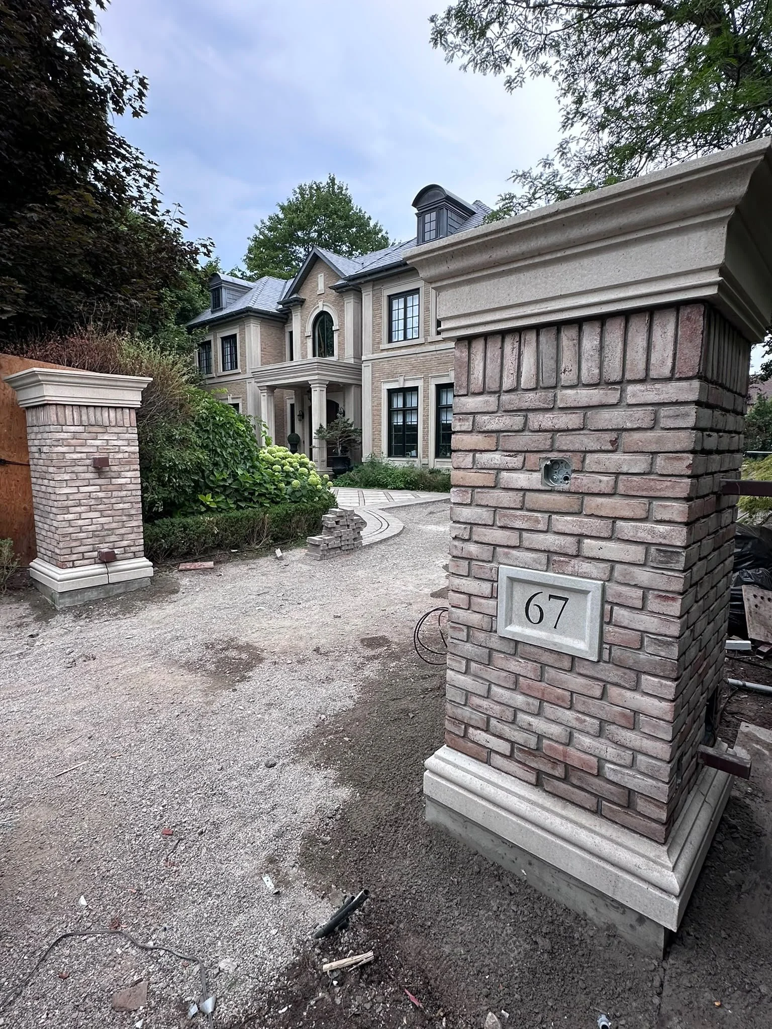 A brick gate pillar with the number 67 on it, part of a wall under construction in front of a large house with a brick and stone facade, multiple windows, and a front porch, surrounded by trees.