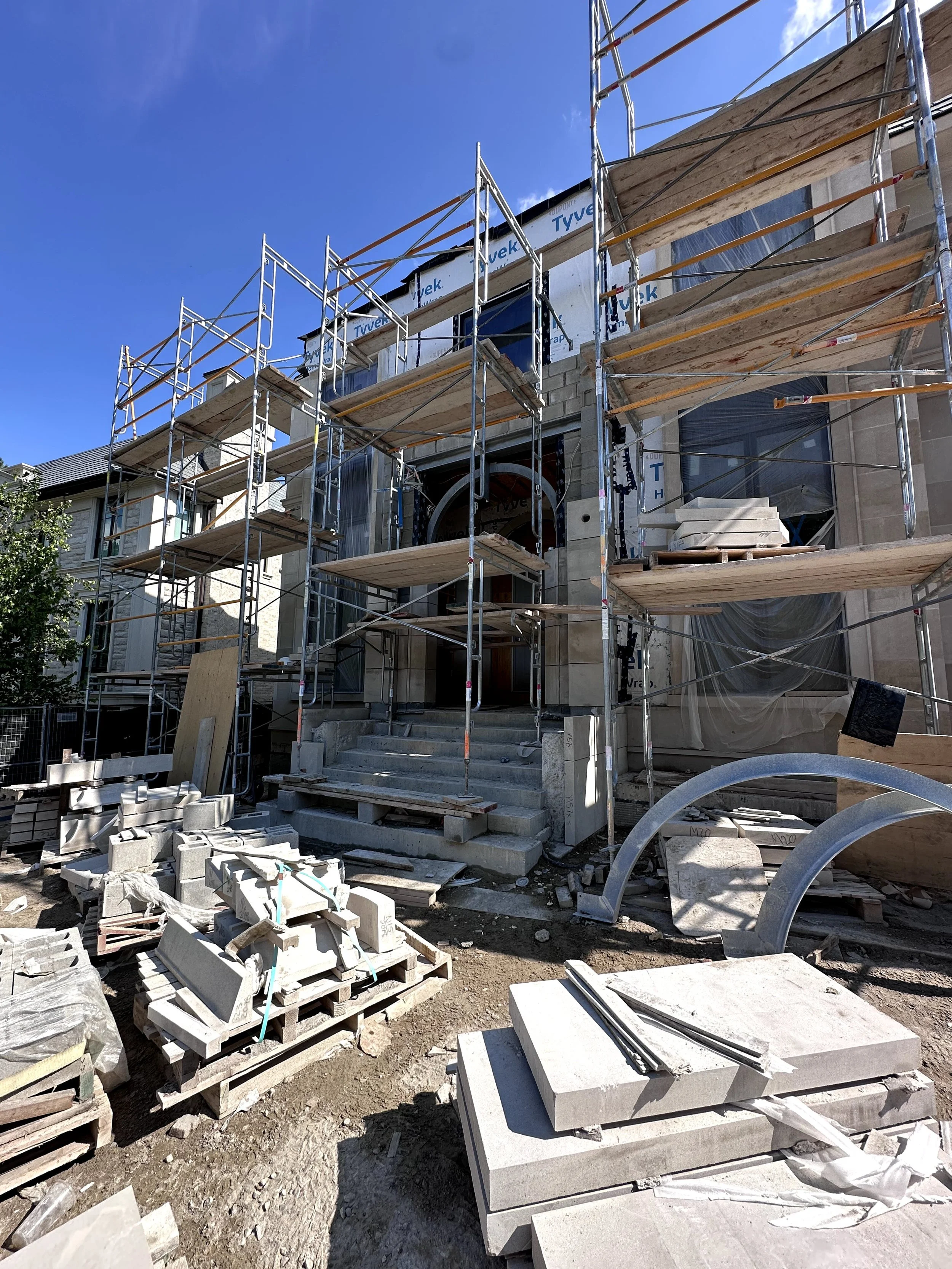 Construction site of a house with scaffolding around it. The house has a stone facade and large windows, with building materials and debris scattered on the ground.