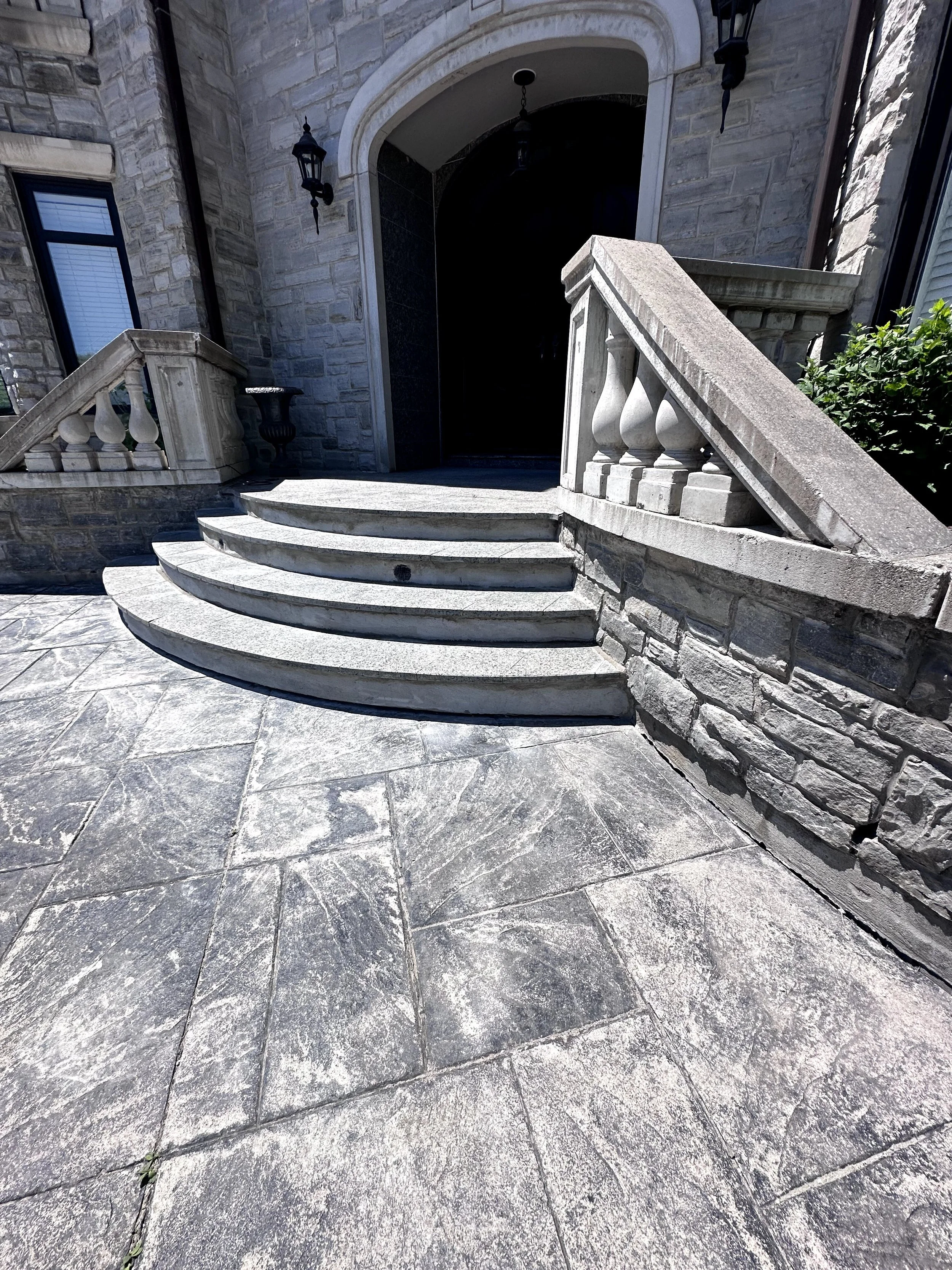 Stone steps leading up to an arched doorway on a brick house with black lantern-style wall lights and a small black planter on the side. The ground is paved with stone tiles.