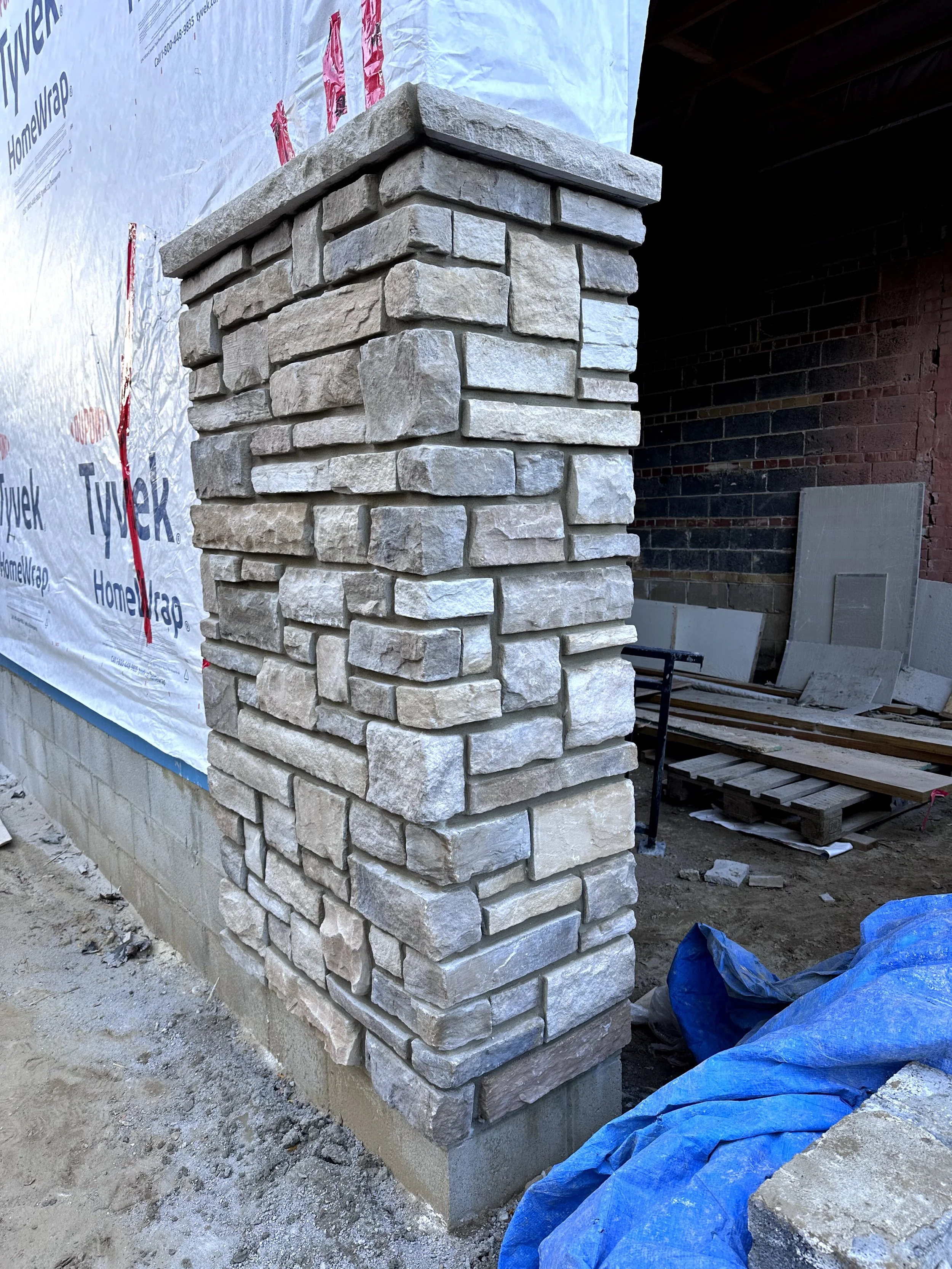 Construction site featuring a stone brick column with white stones and a concrete cap, with building materials and a blue tarp surrounding the base.