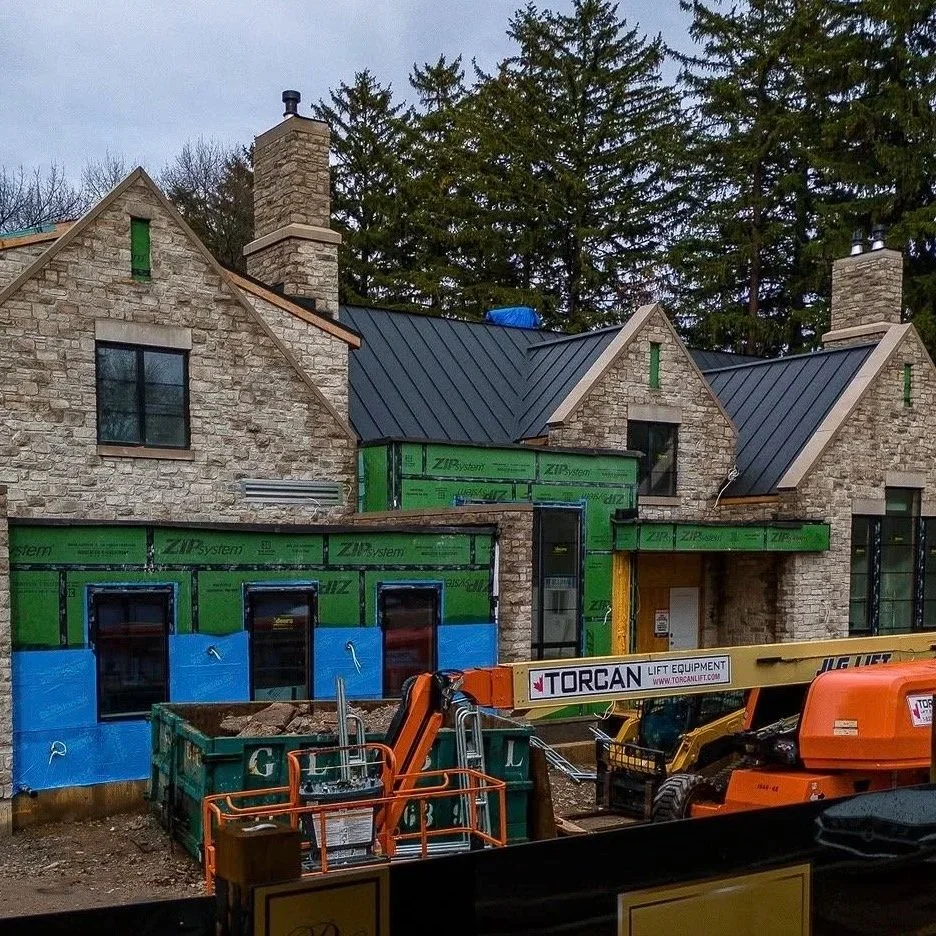Under construction of a stone house with a black metal roof, green sheathing, and large chimney, with construction machinery and equipment in the foreground.