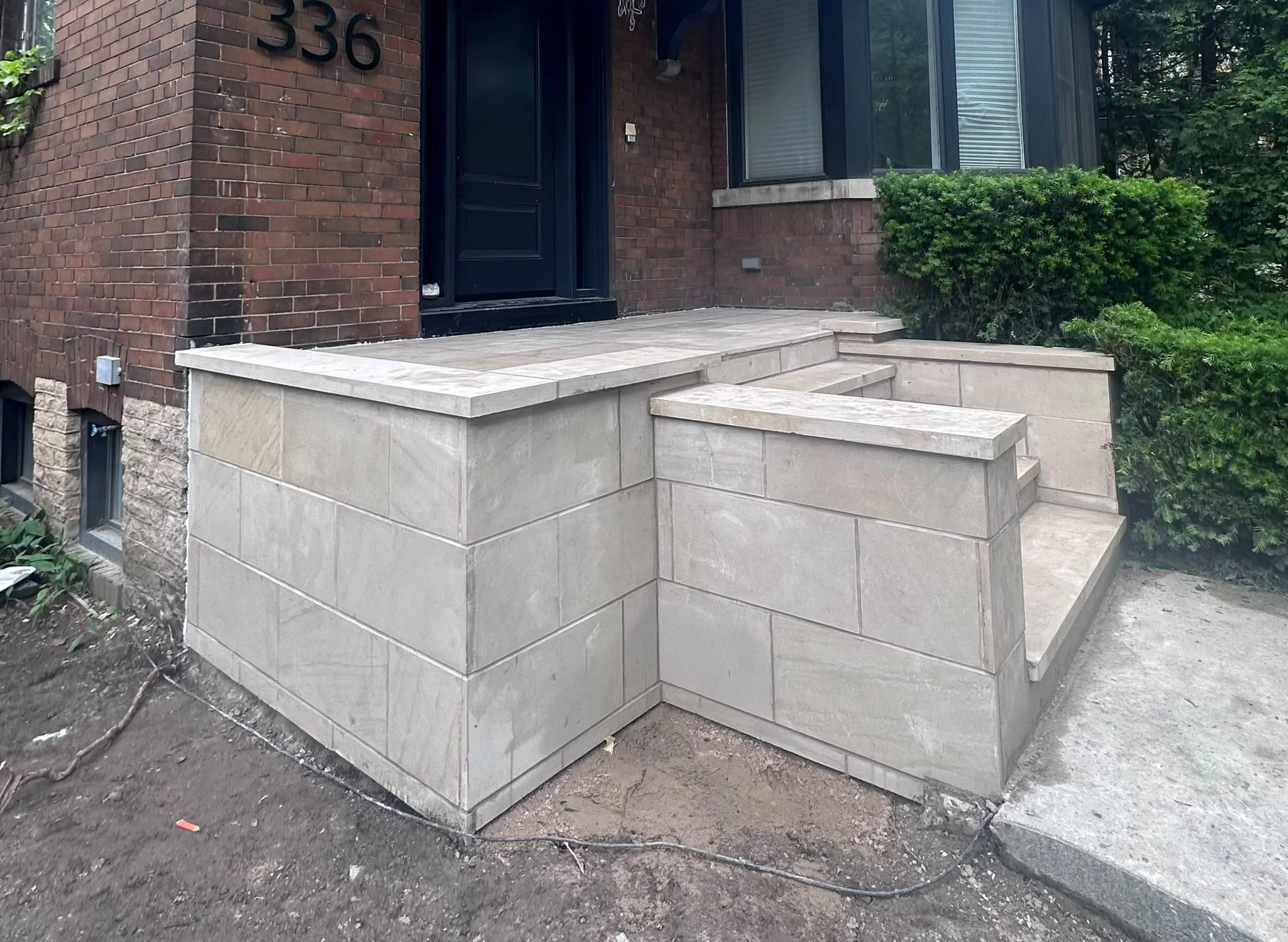 Front porch area with freshly installed light-colored stone steps and platform, brick exterior wall with house number 336, a black door, a window with blinds, and shrubs nearby, with dirt ground in the foreground.