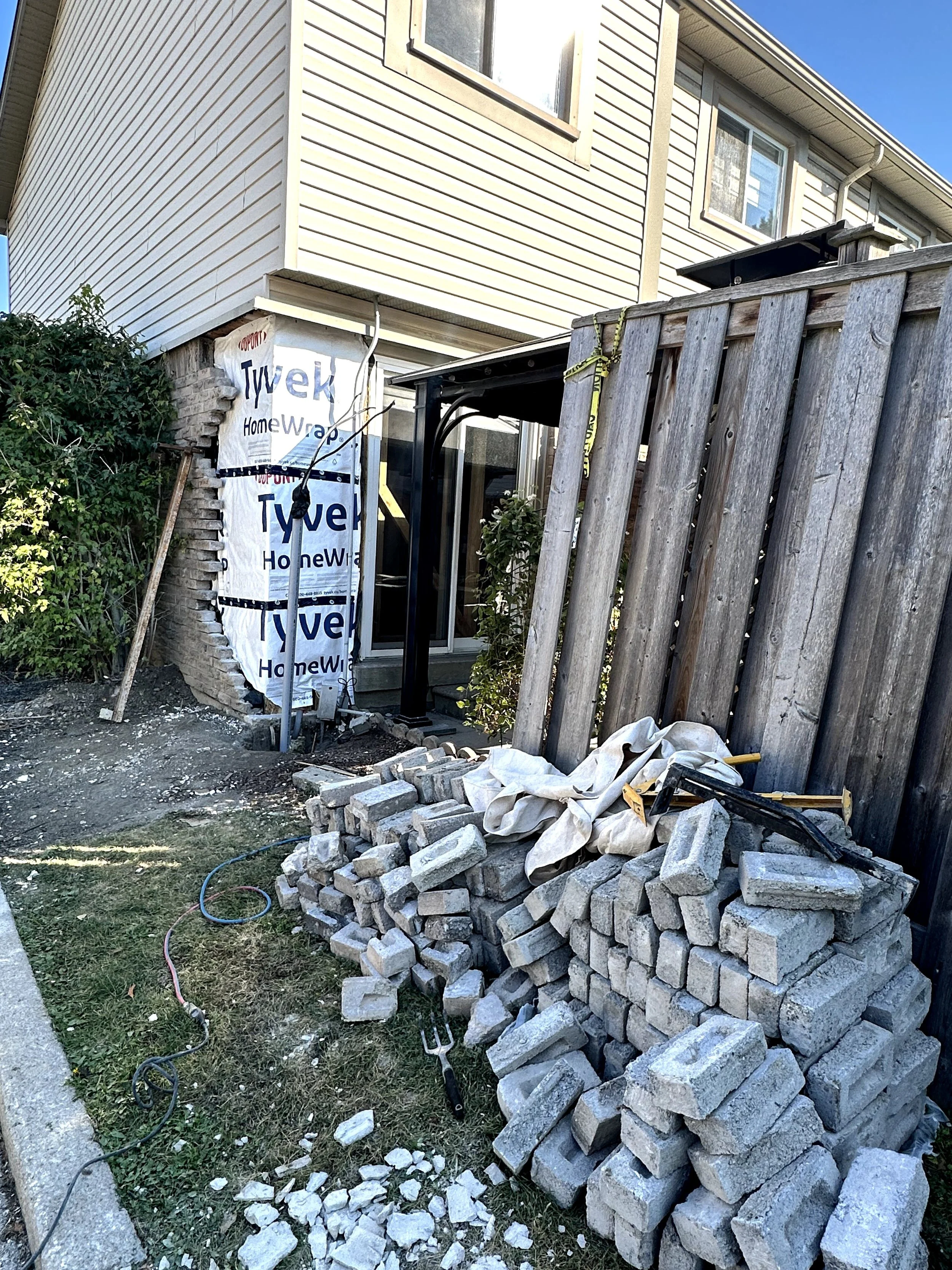 Construction site with a pile of gray bricks, some scattered on the ground. A wooden fence and the exterior of a house with beige siding, windows, and a small porch can be seen. Construction tools and materials are present, including a cloth, a garde
