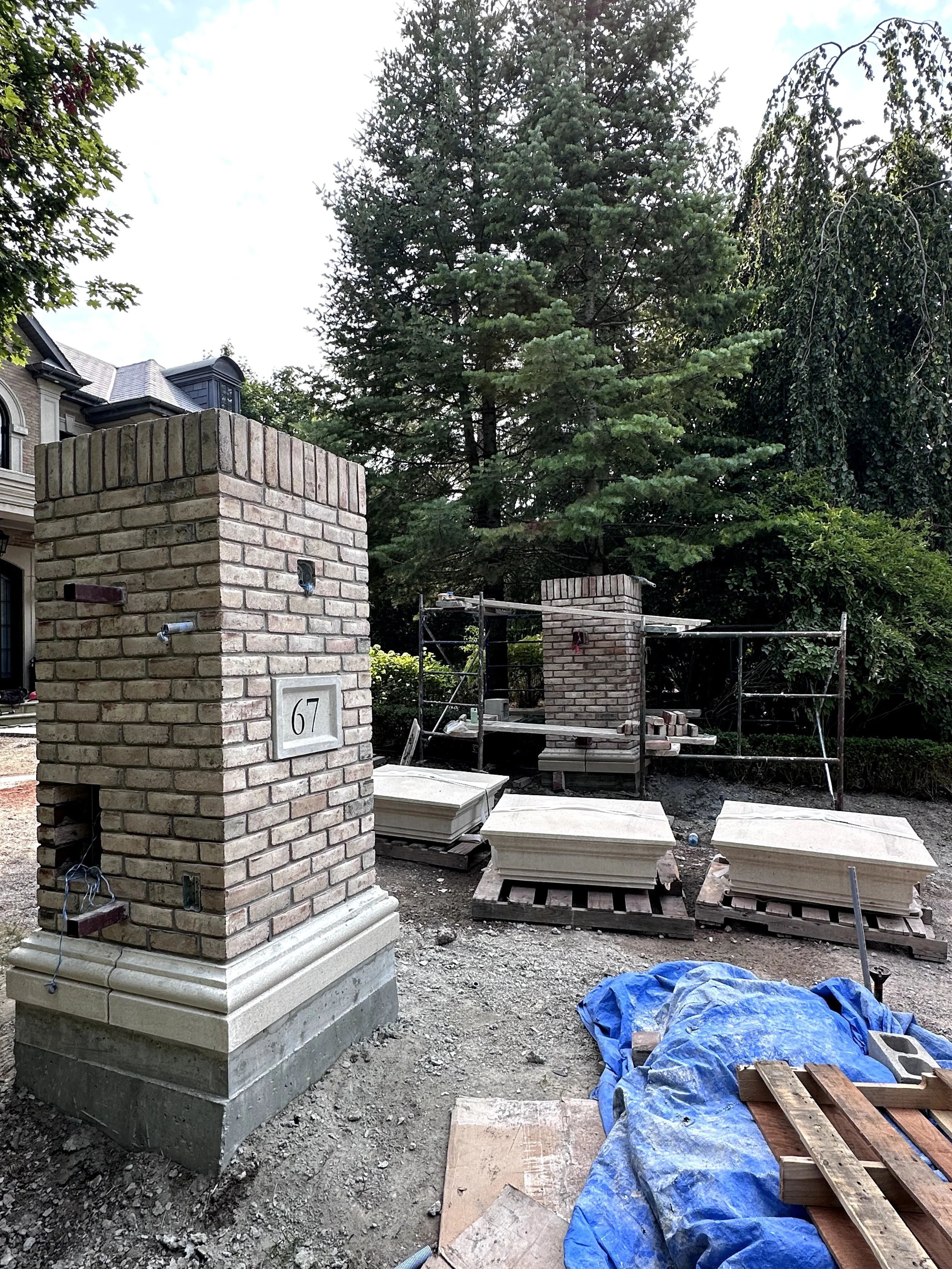 Construction site with brick columns, construction materials, and scaffolding, surrounded by trees and a blue tarp on the ground.