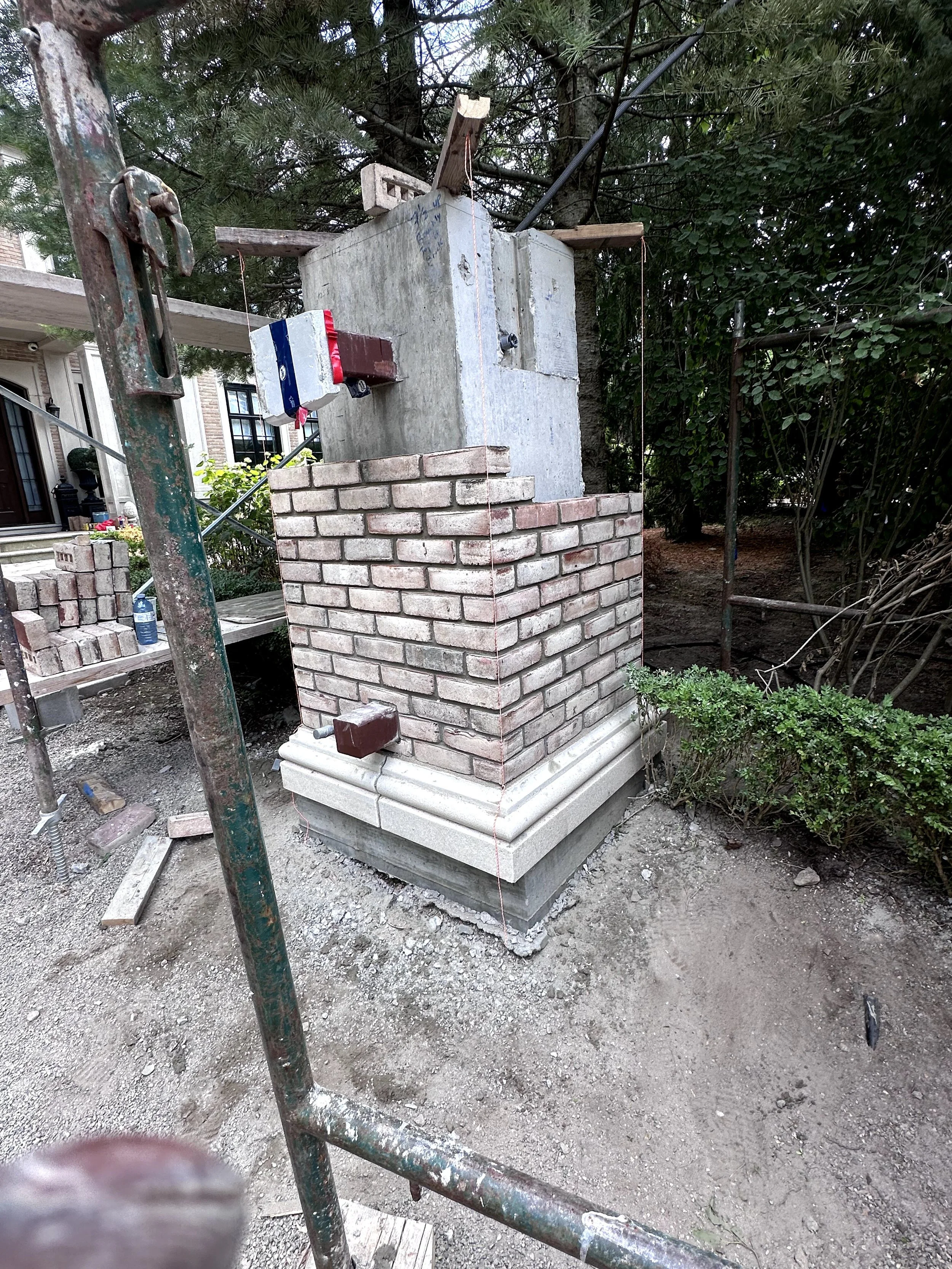 A construction site showing a partially built brick pillar with a concrete base, surrounded by scaffolding and building materials, with trees and a house visible in the background.