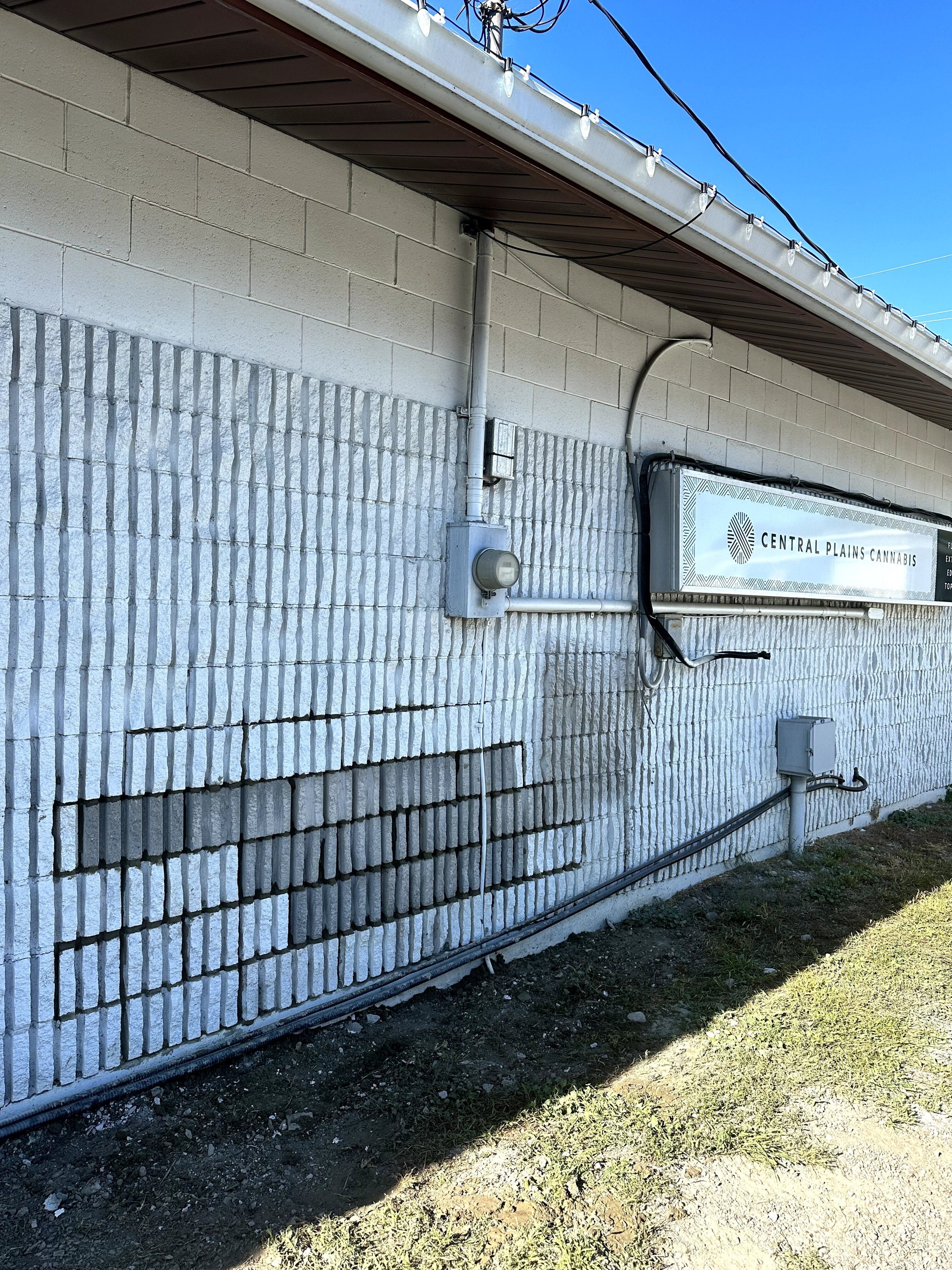 The image shows the exterior of a building with a white brick wall. There is a sign that reads 'CENTRAL PLAINS CANNABIS' mounted on the wall. Electrical wires and conduit are visible, along with some outdoor lighting fixtures. The area in front of th