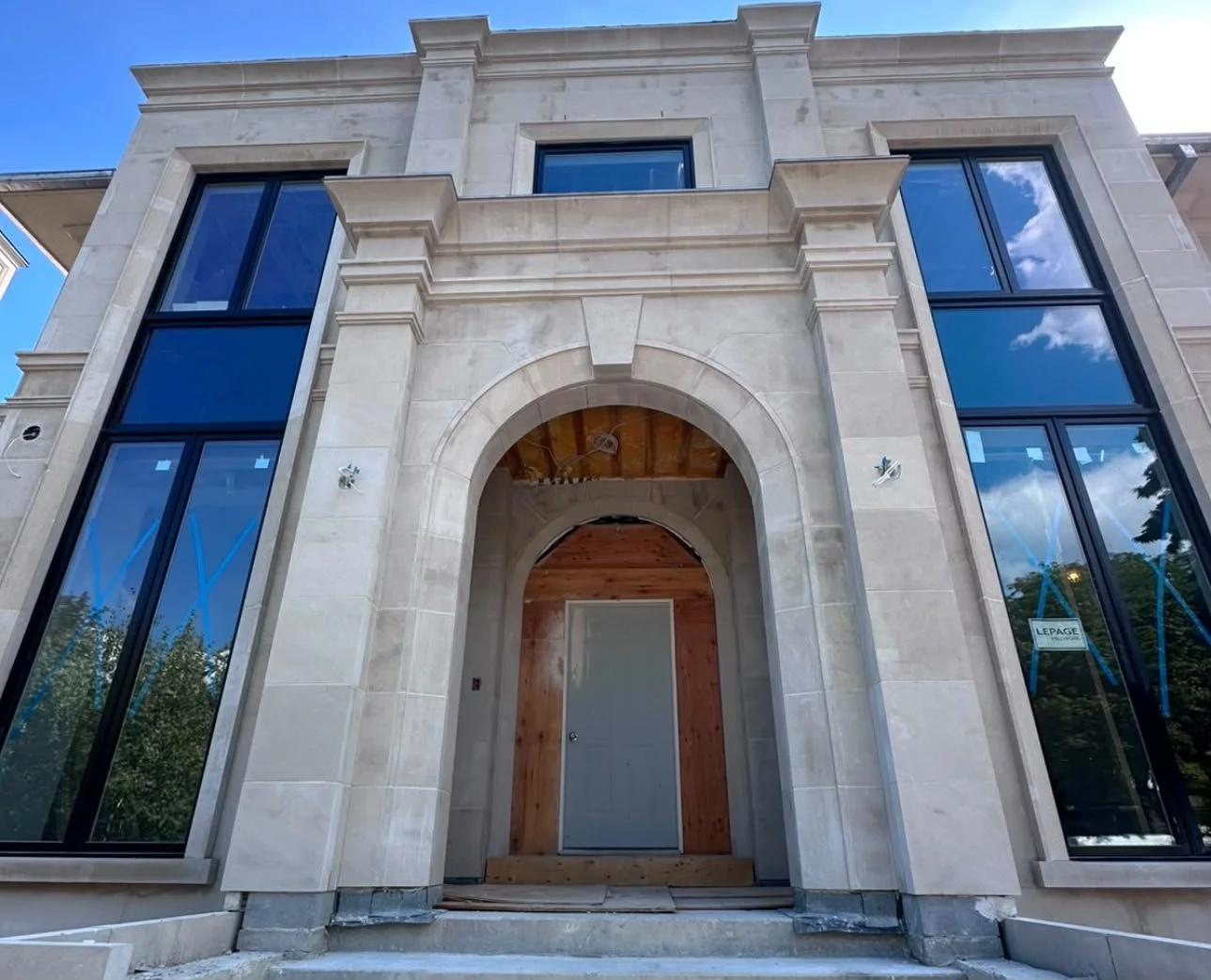 Front view of a modern, unfinished residential building with large windows and an arched entryway, under construction, with some exposed wood and concrete.