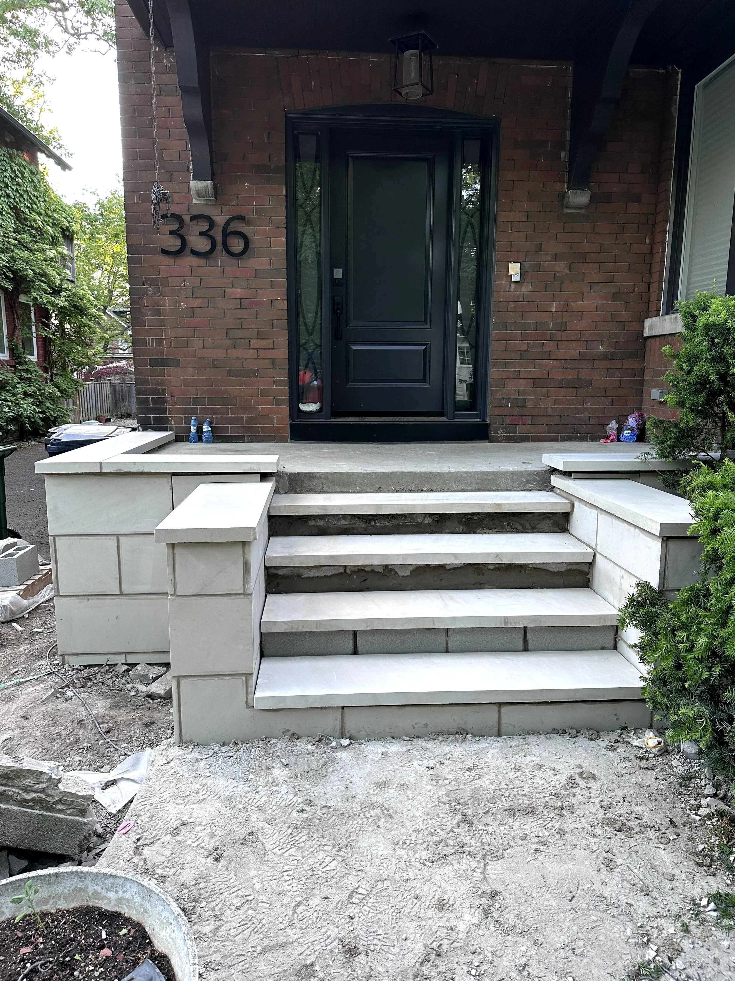 Front porch of a brick house with a black door, steps, and temporary construction work. Two water bottles are on the steps, with some construction materials and tools scattered on the ground.
