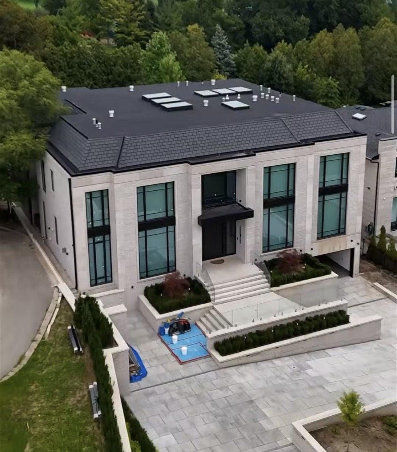 An aerial view of a modern multi-story house with large glass windows, a black roof with skylights, and landscaped surroundings including a patio and green trees in the background.