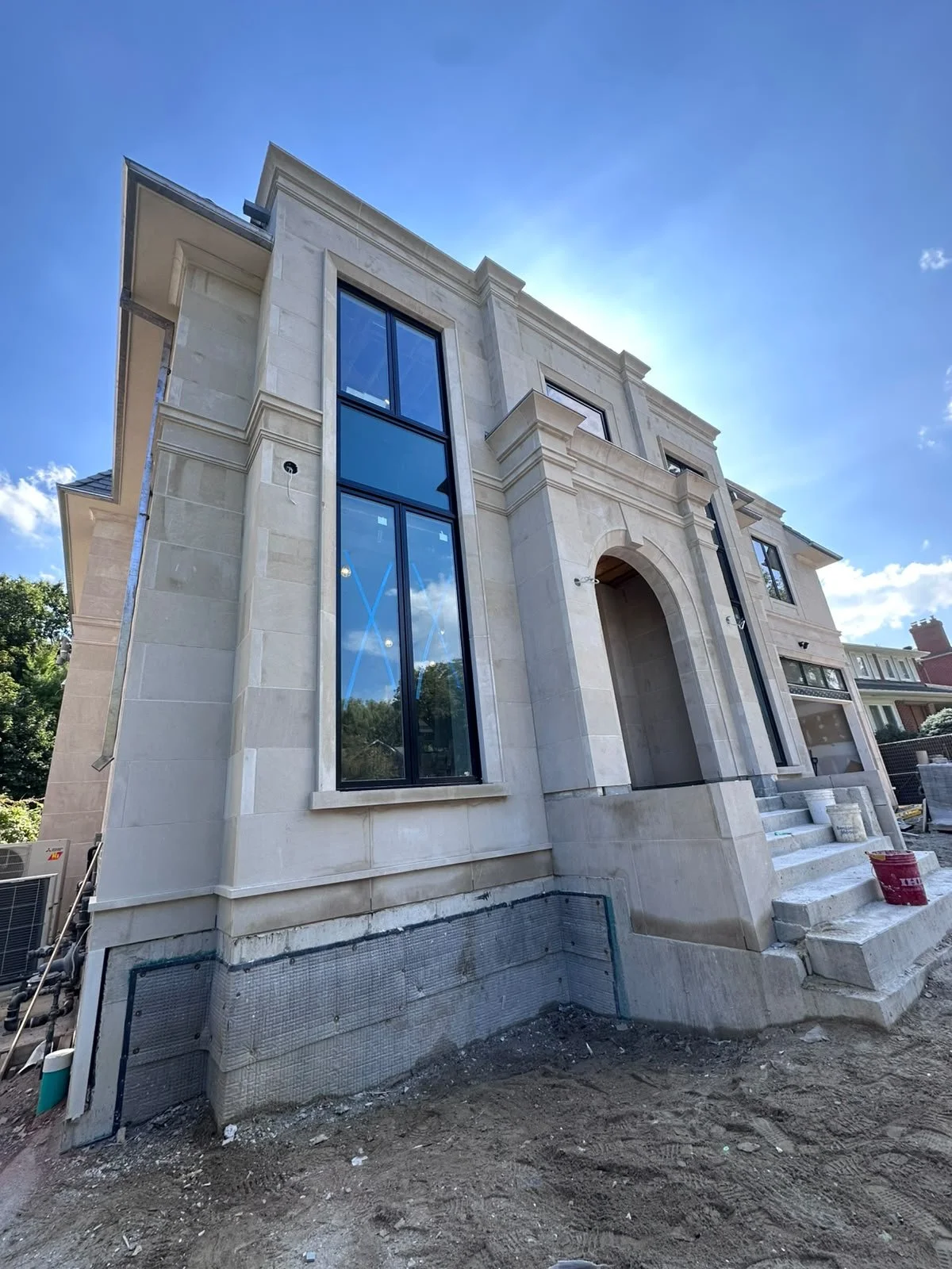 A large, modern house under construction with a stone facade, tall windows, and steps leading up to the front entrance, with construction materials on the ground.