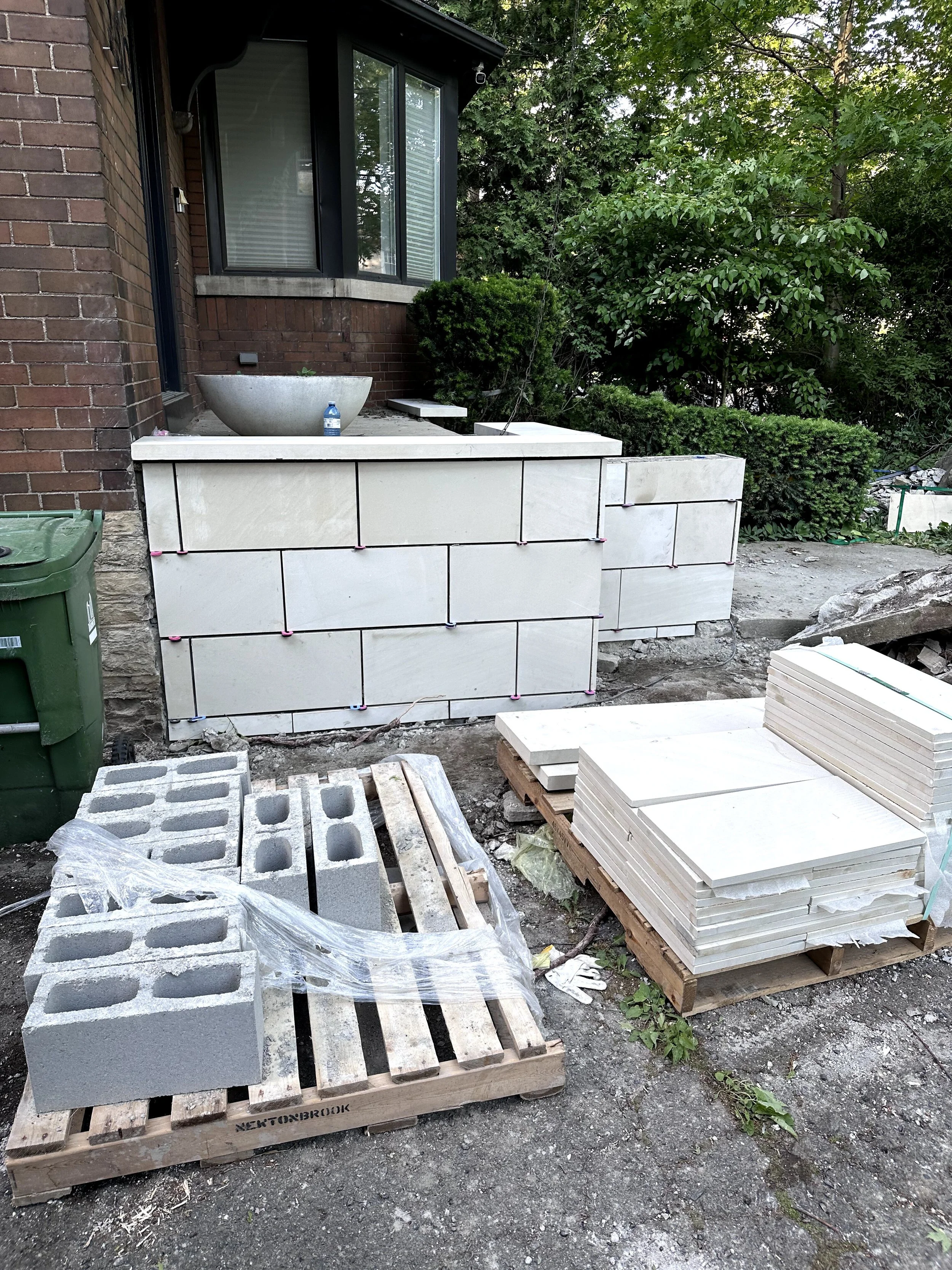 Construction site with white tiles stacked on a wooden pallet, cinder blocks, and a partially built outdoor structure with a brick wall and a window, surrounded by greenery.