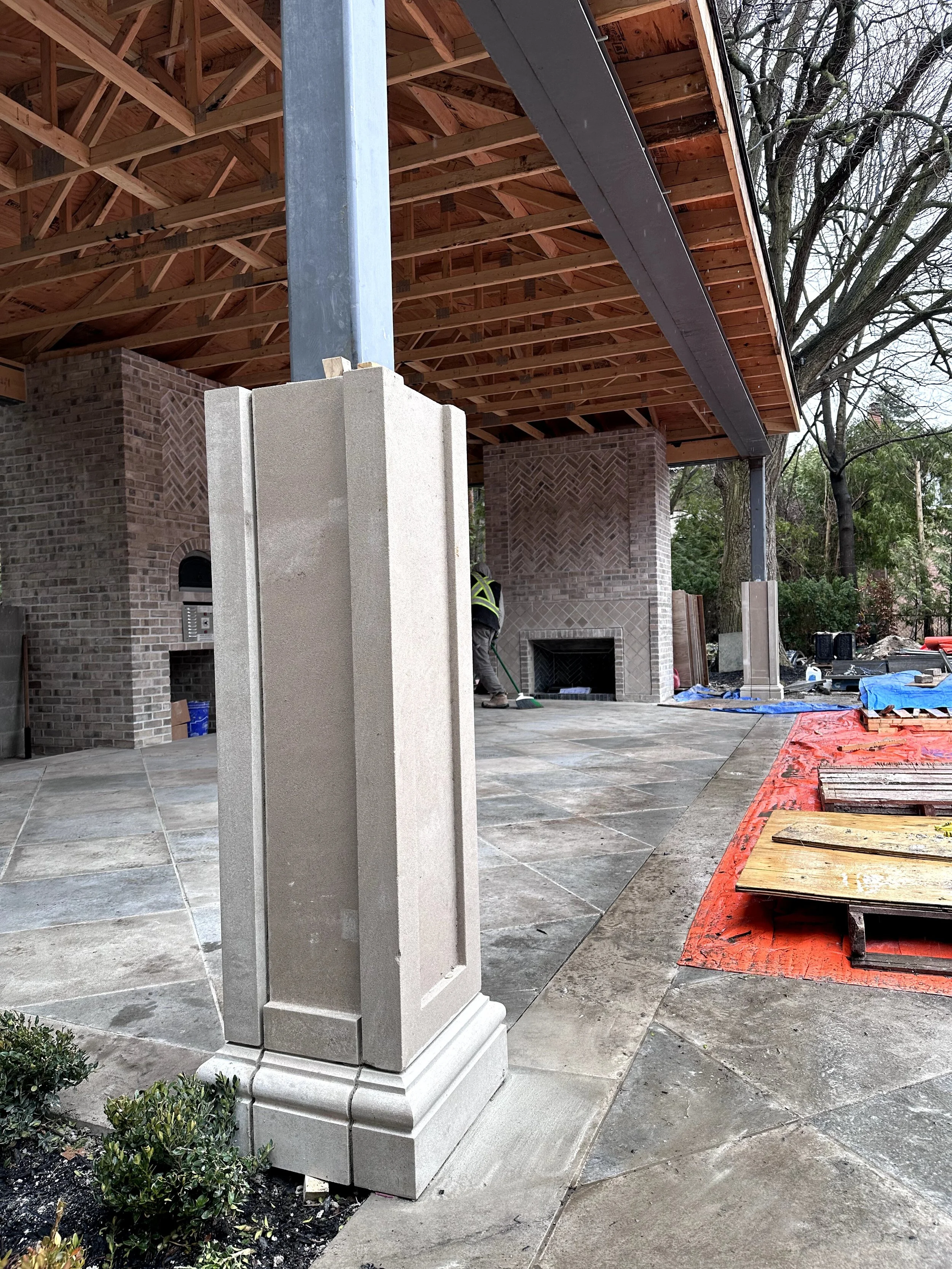 Construction site showing a partially built patio or porch with wooden roofing, brick fireplace, and decorative stone column.