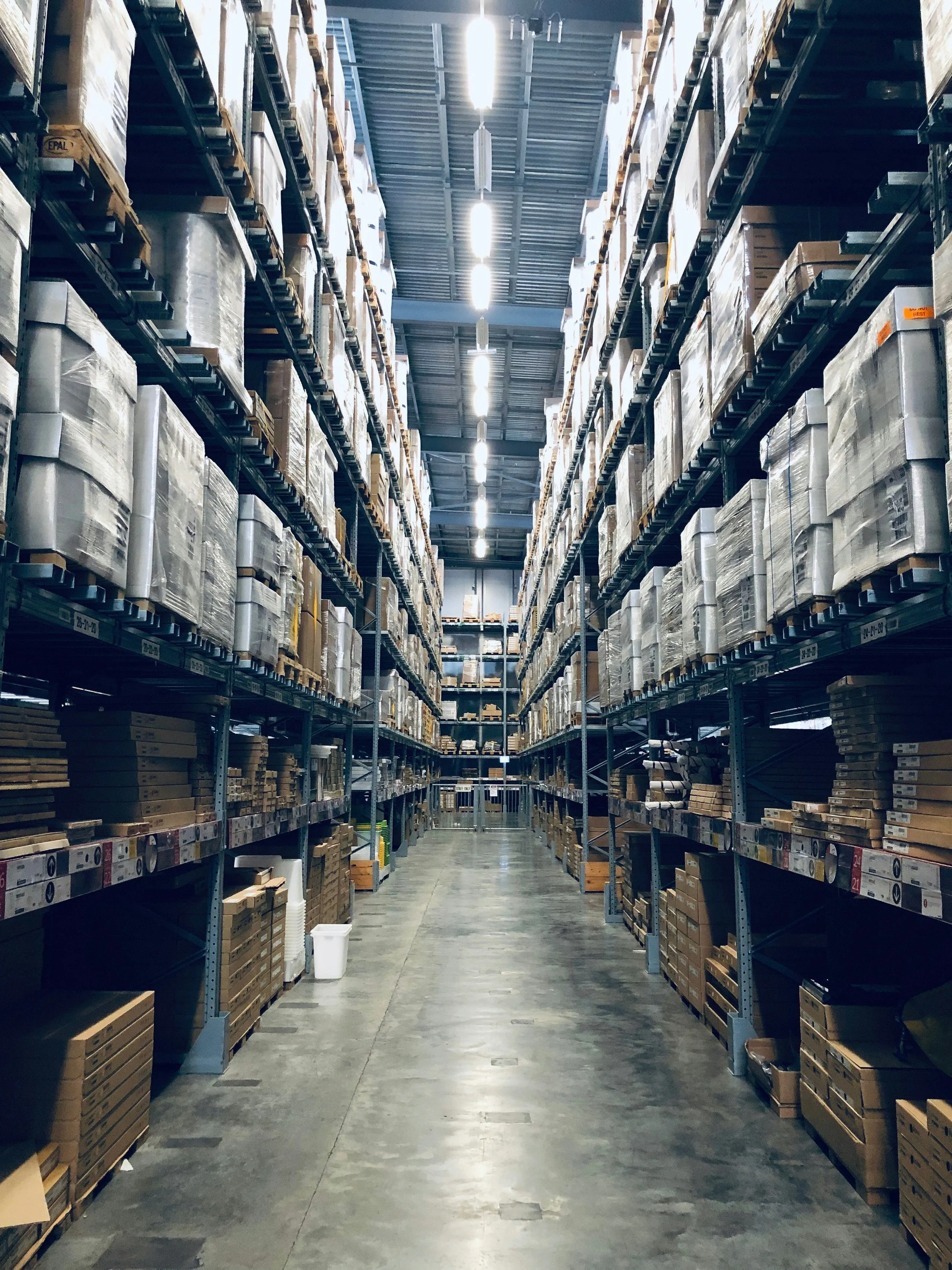 View of tall warehouse shelves filled with cardboard boxes and packaging items in an aisle with overhead lights. The shelves are organized with labels underneath each section.