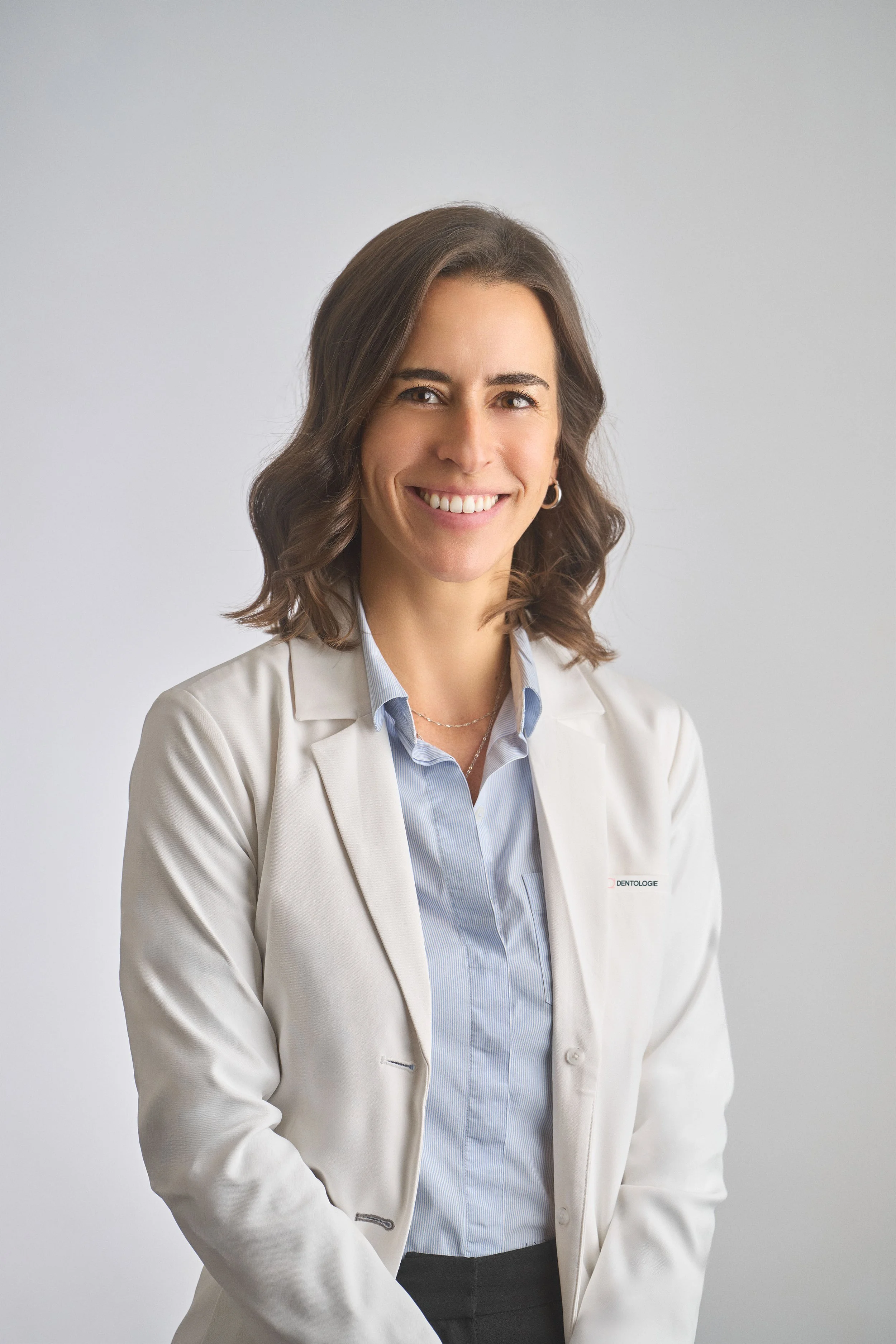 Portrait d’une dentiste souriante portant une blouse blanche, devant un fond gris clair.