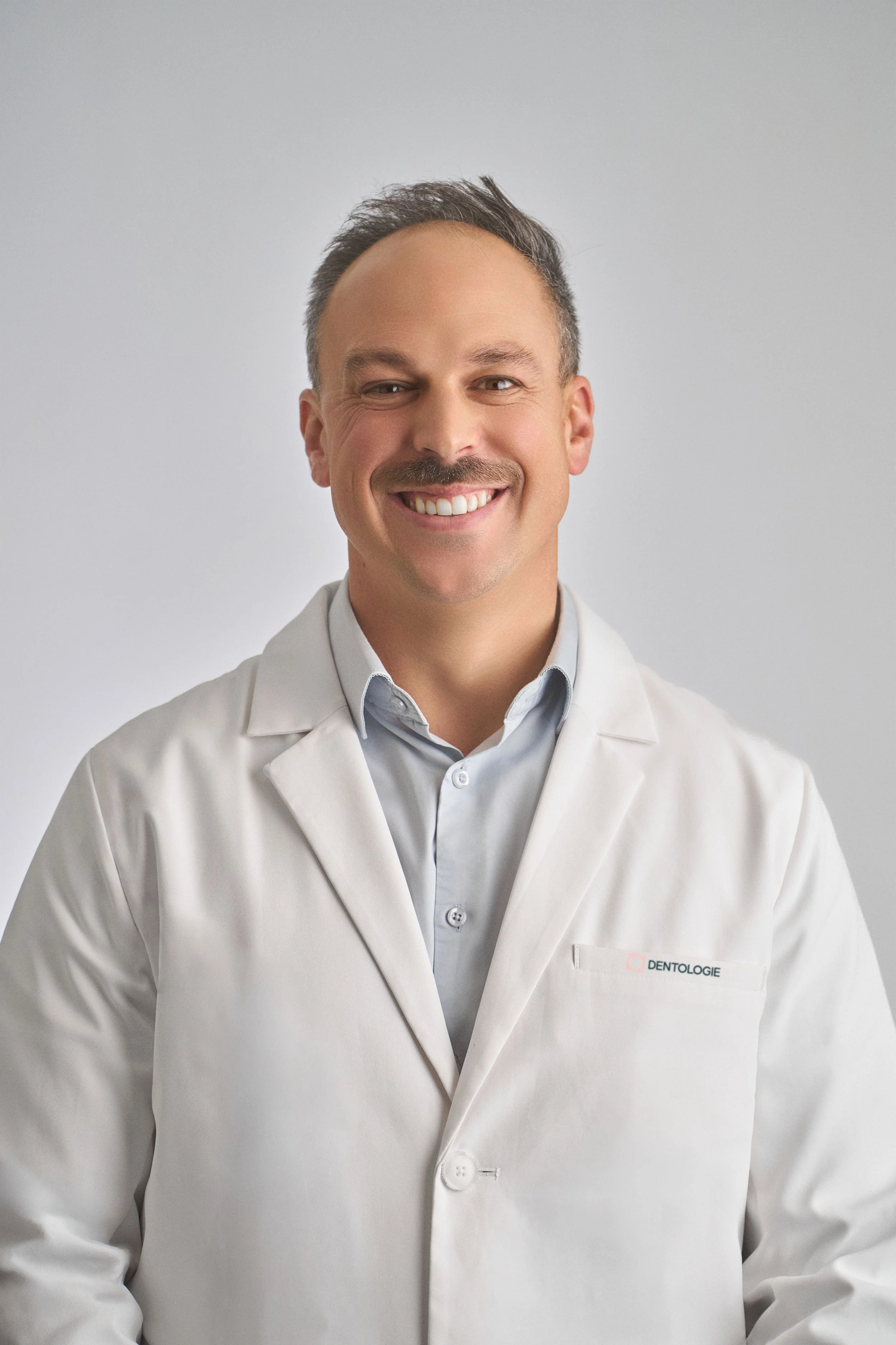 Portrait d’un dentiste souriant portant une blouse blanche avec le logo « Dentologie ».