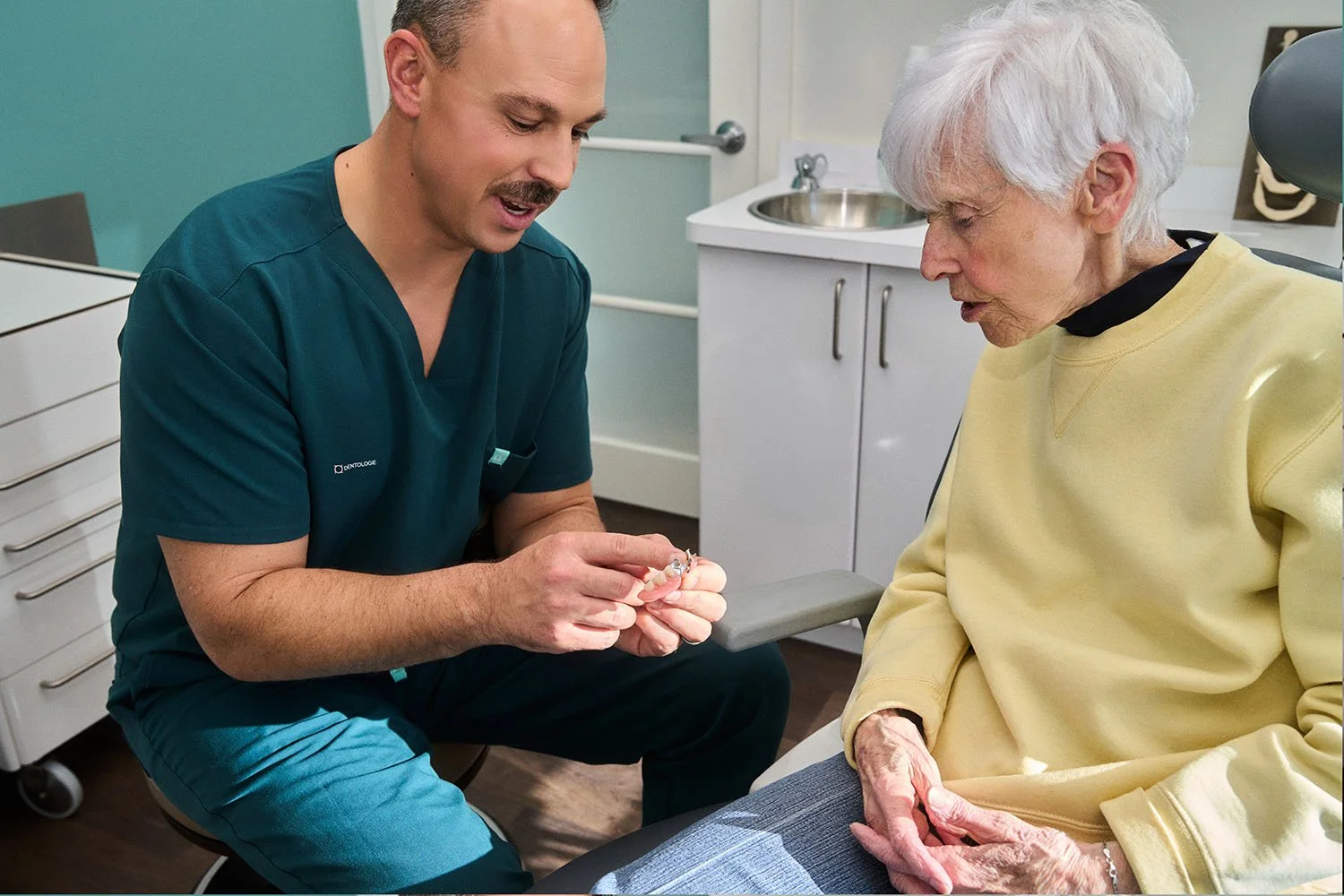 Professionnel de santé montrant une prothèse dentaire à une patiente âgée dans un cabinet médical.