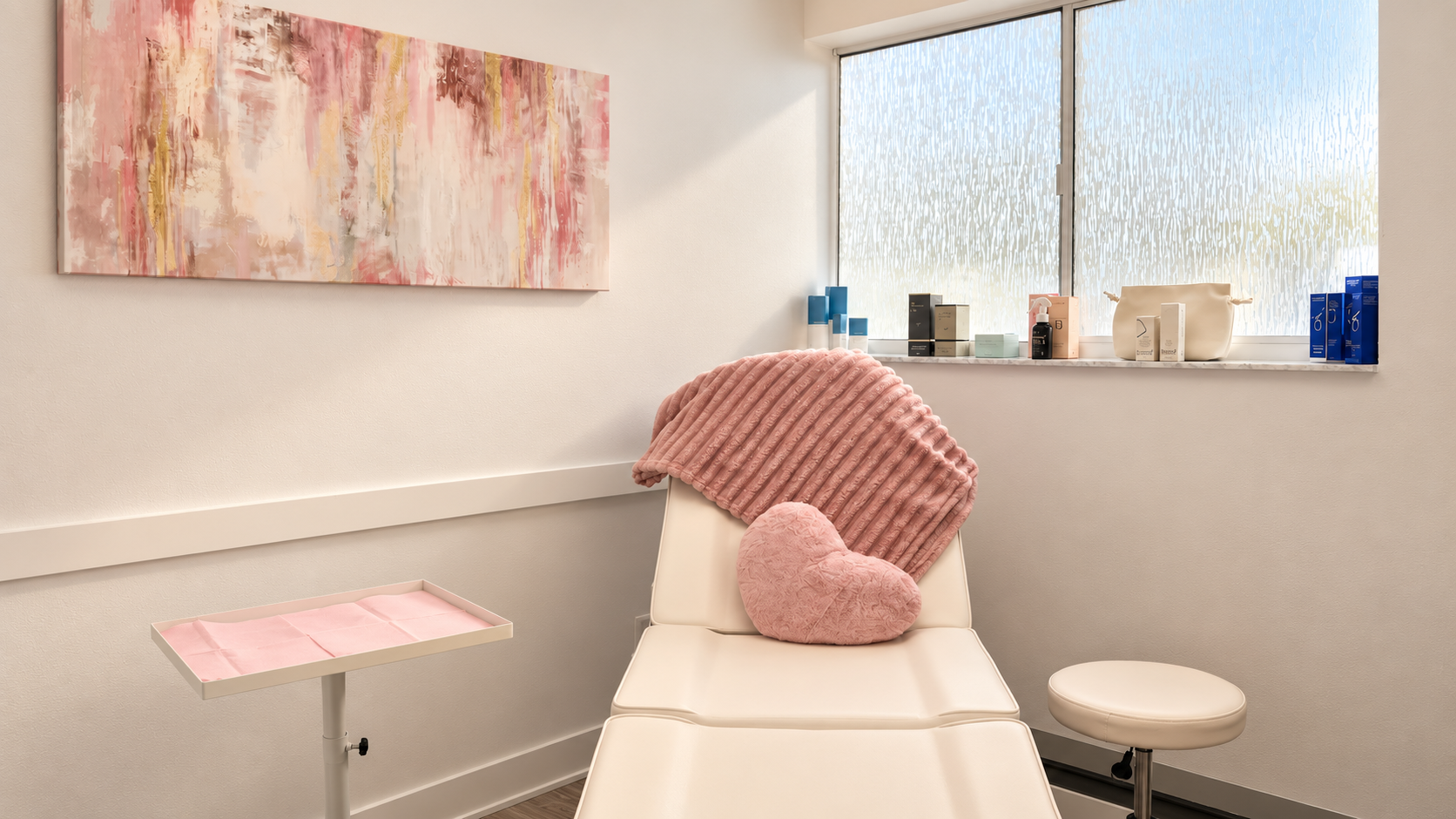 A medical or consultation room with a white examination chair draped with a pink, plush throw blanket and a heart-shaped pink pillow. There is a pink tray on a pole to the left, and a white stool on wheels to the right. A large abstract painting with pink, white, and gold tones hangs on the wall, and a window with frosted glass allows natural light in, with various small product boxes and bottles on the windowsill.