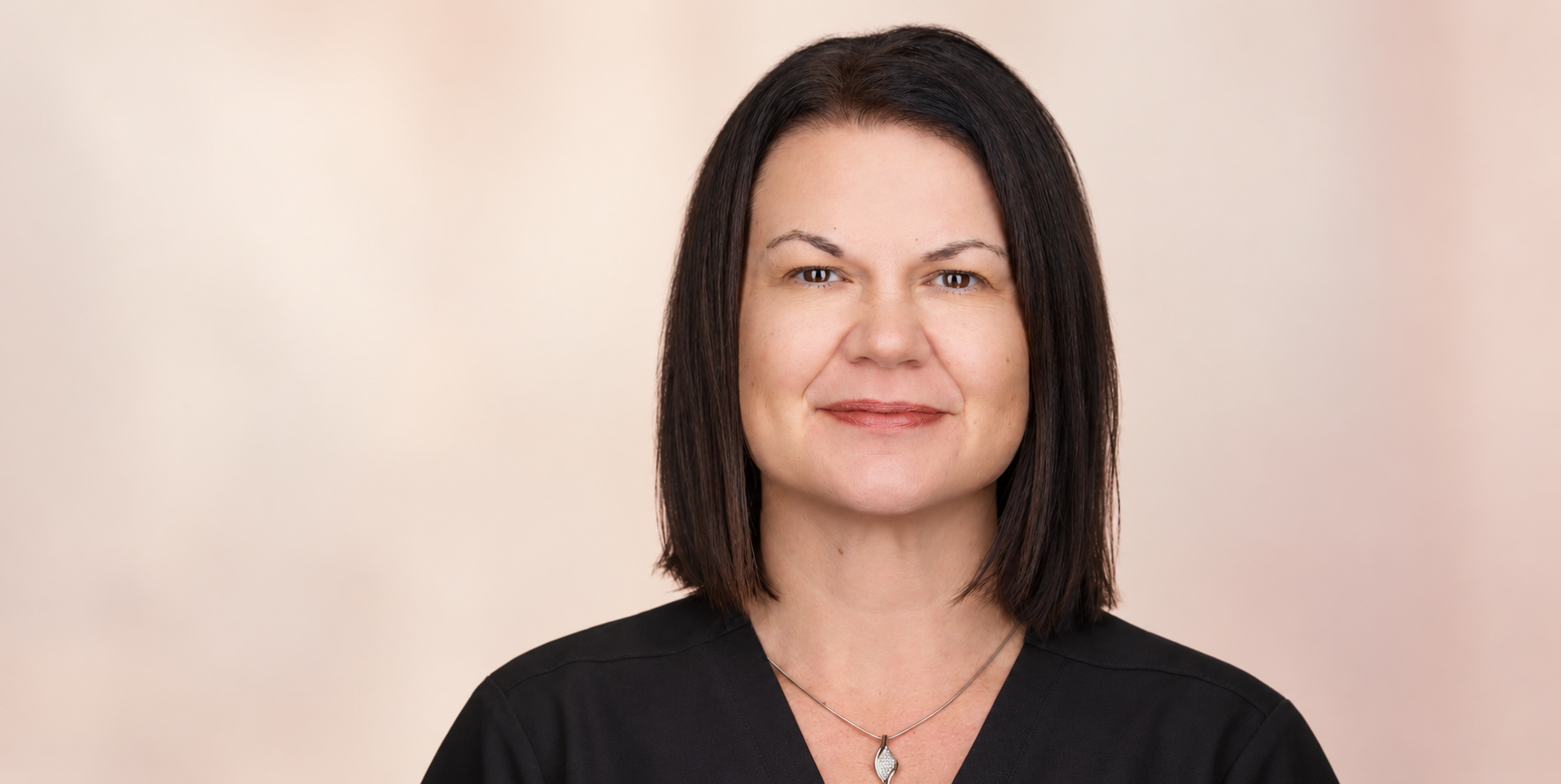 A woman with dark shoulder-length hair, wearing a black top and a silver necklace, smiling softly against a neutral background.