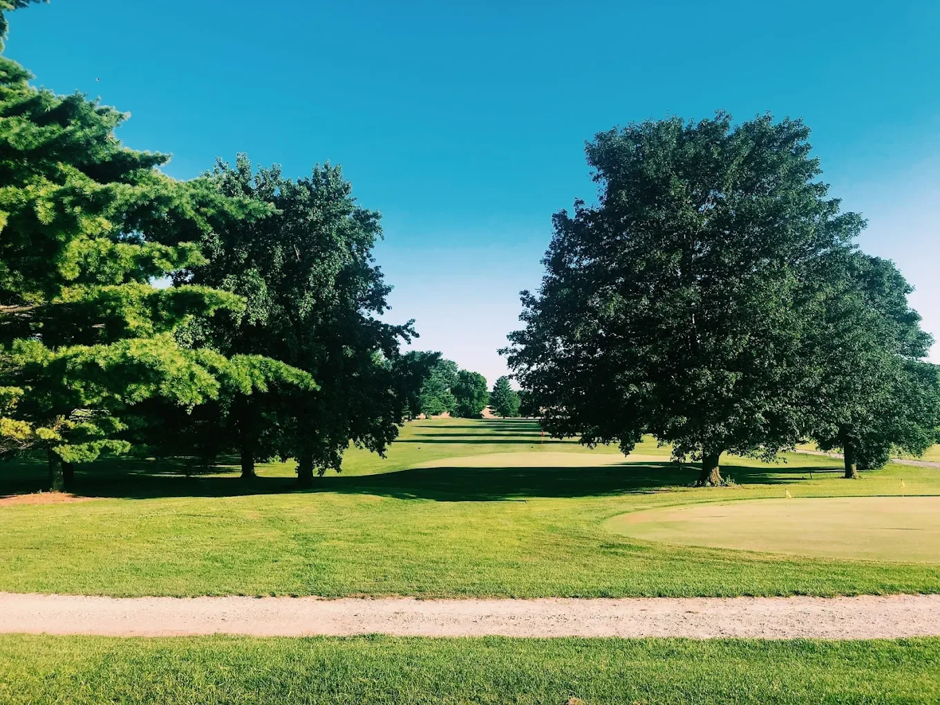 A golf course with green grass, trees, and a sand trap, under a clear blue sky.