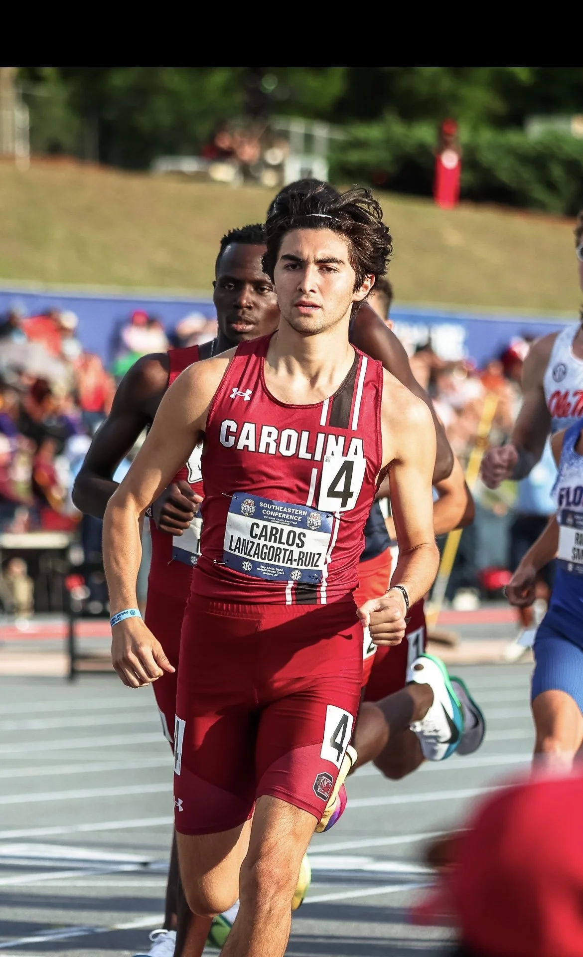 A male marathon runner wearing a red uniform with 'Carolina' written on it, competing in a race. The runner's bib shows "Carlos Lanzagorta-Ruiz." He has dark hair and a focused expression. Other runners and spectators are visible in the background.