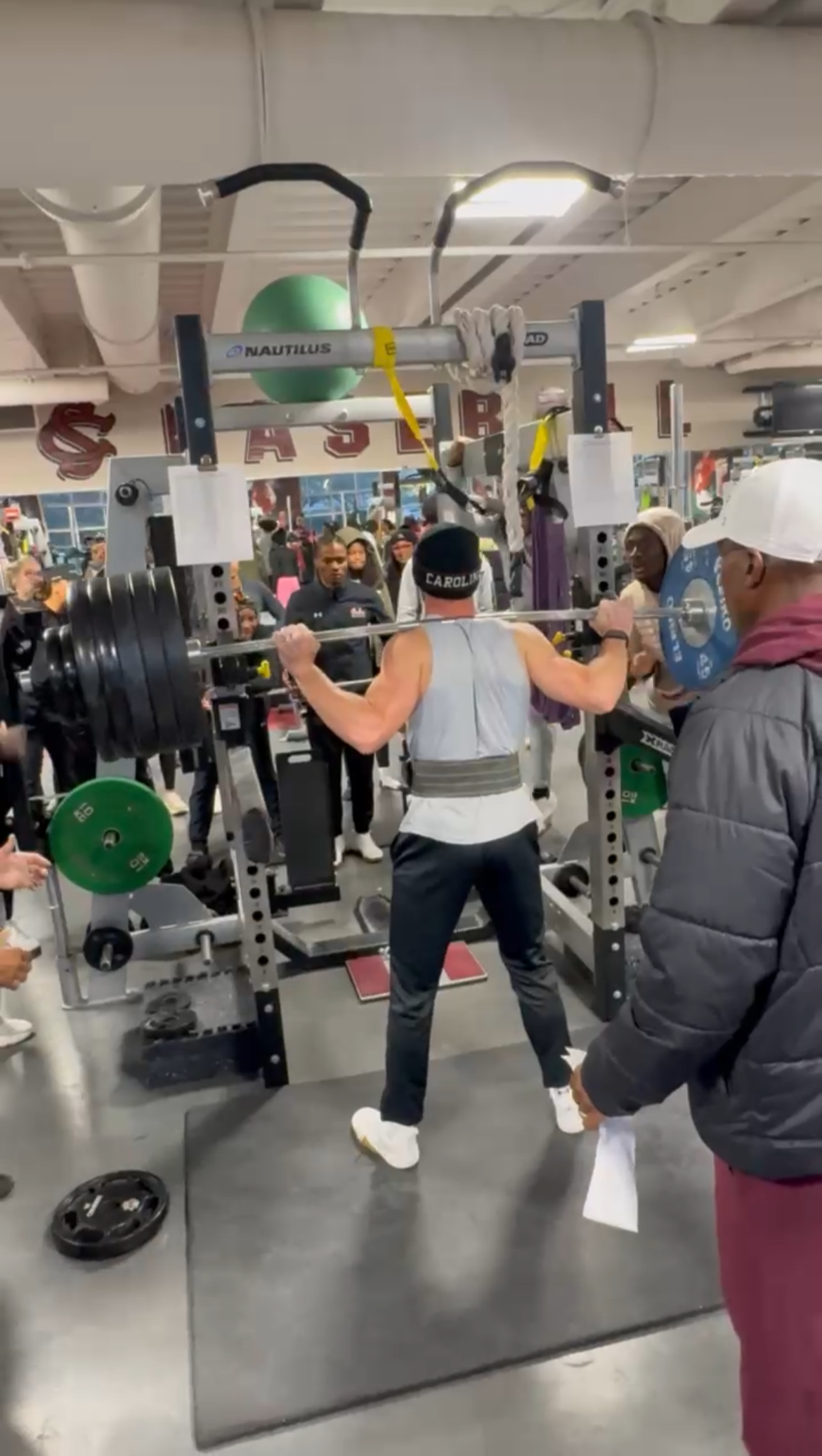 A person lifting a barbell in a gym surrounded by people watching, gym equipment visible, including plates and a green exercise ball.